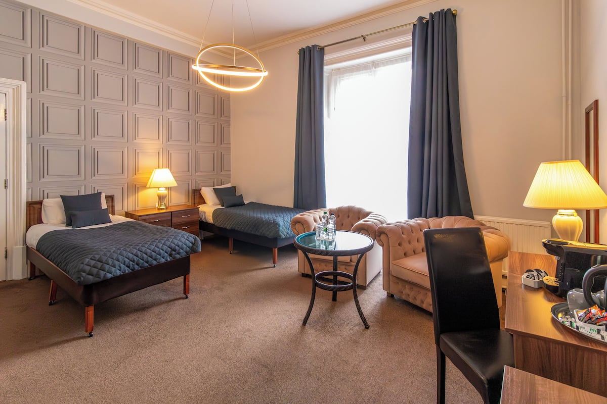 Ein Hotelzimmer mit einem Bett, zwei Nachttischen, einem kleinen runden Tisch, einem Sofa, einem Sessel und einem Schreibtisch mit einer Lampe.