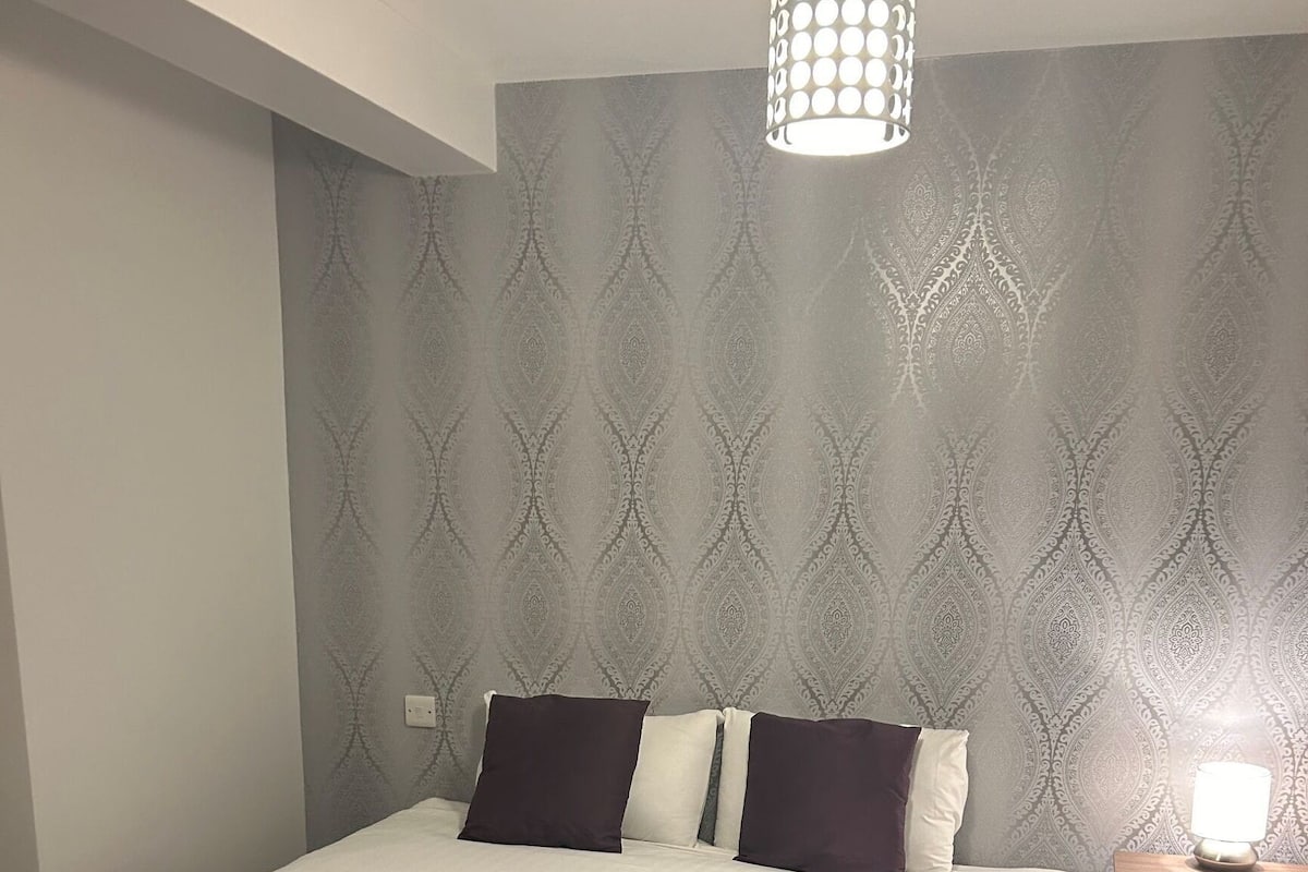 Ein ordentlich gemachtes Bett mit einer dunkel gefärbten Decke, einer Wand mit Muster und einer Nachttischlampe.