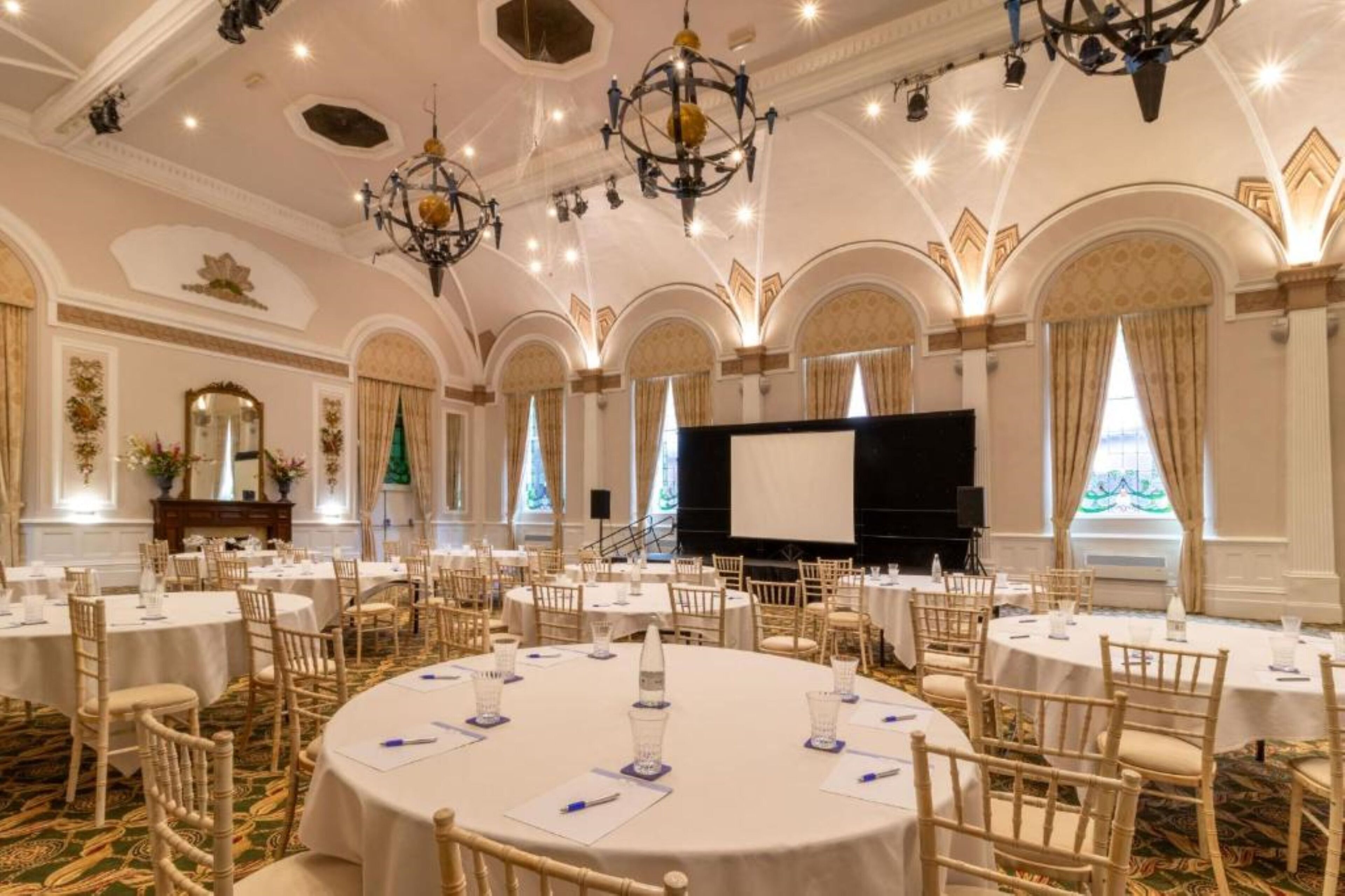 A banquet hall with round tables, chandeliers, and a stage for presentations.