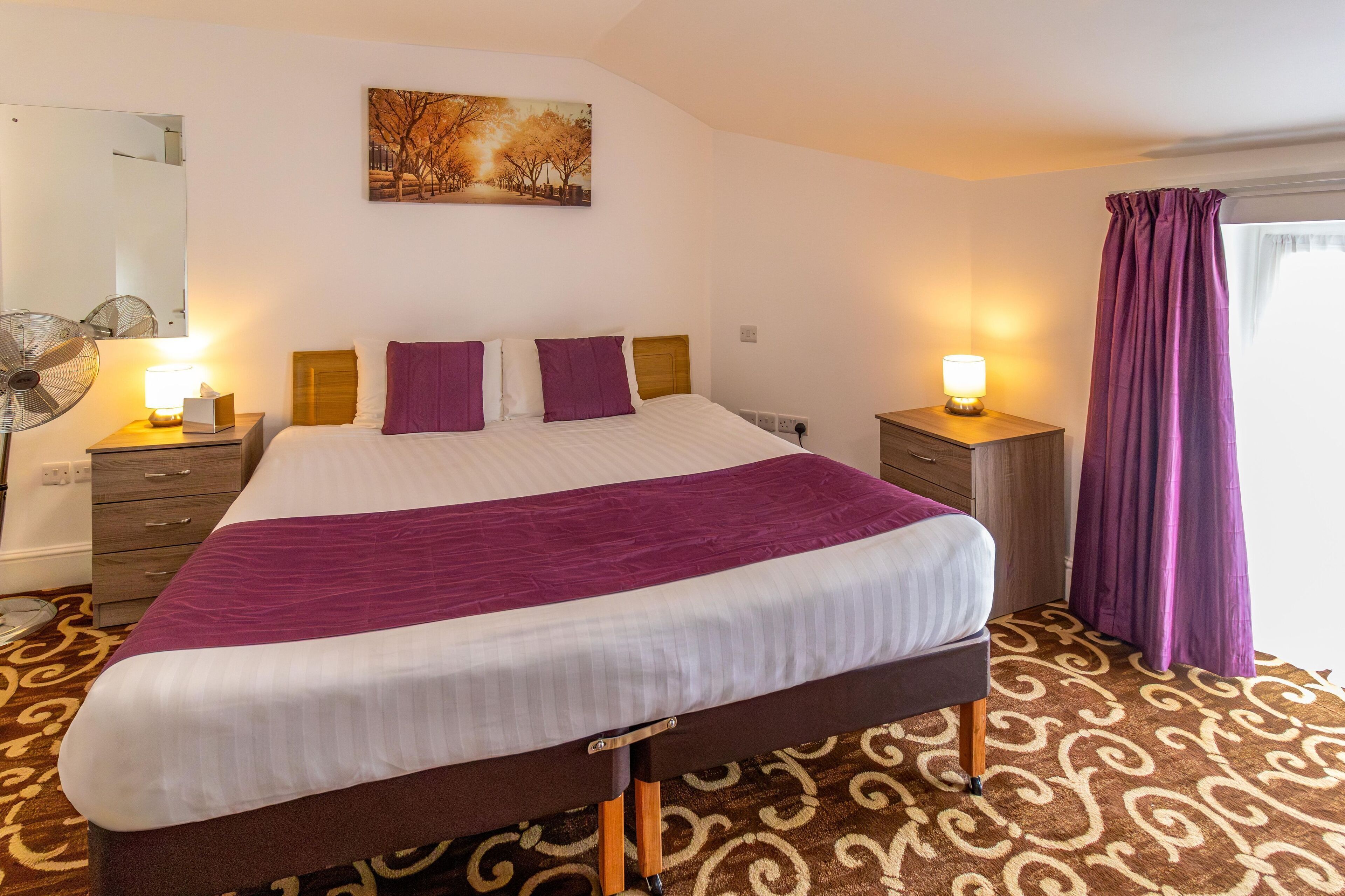 A neatly made bed with a purple blanket, a bedside table with a lamp, and a wall-mounted fan.