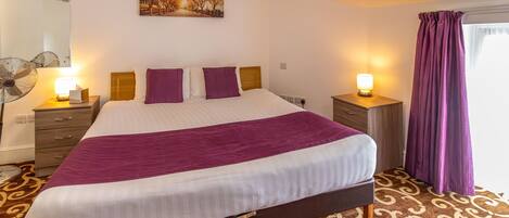 A neatly made bed with a purple blanket, a bedside table with a lamp, and a wall-mounted fan.