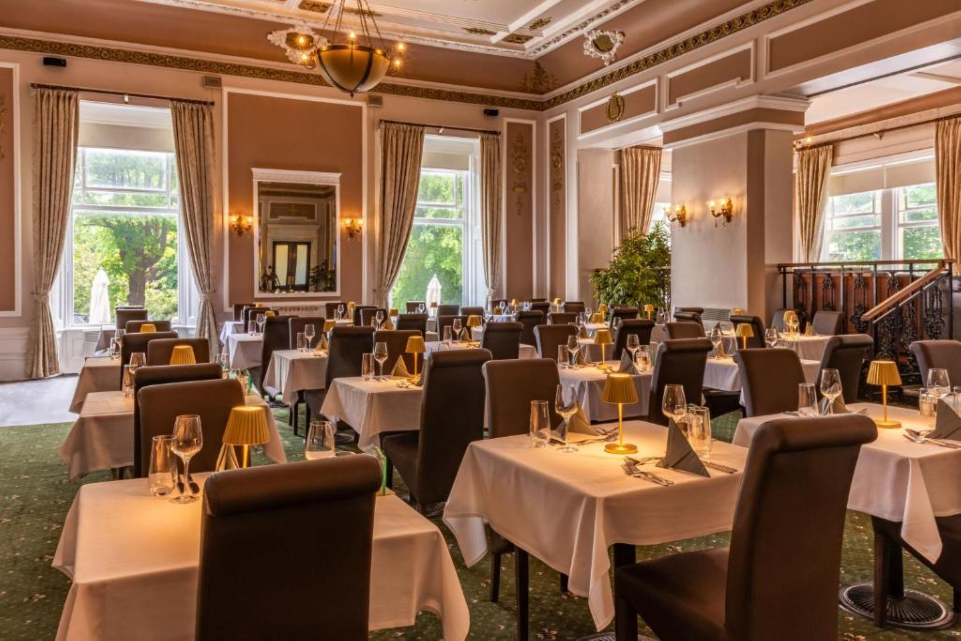 A formal dining room with multiple tables set for guests, large windows with curtains, and a staircase leading to an upper level.