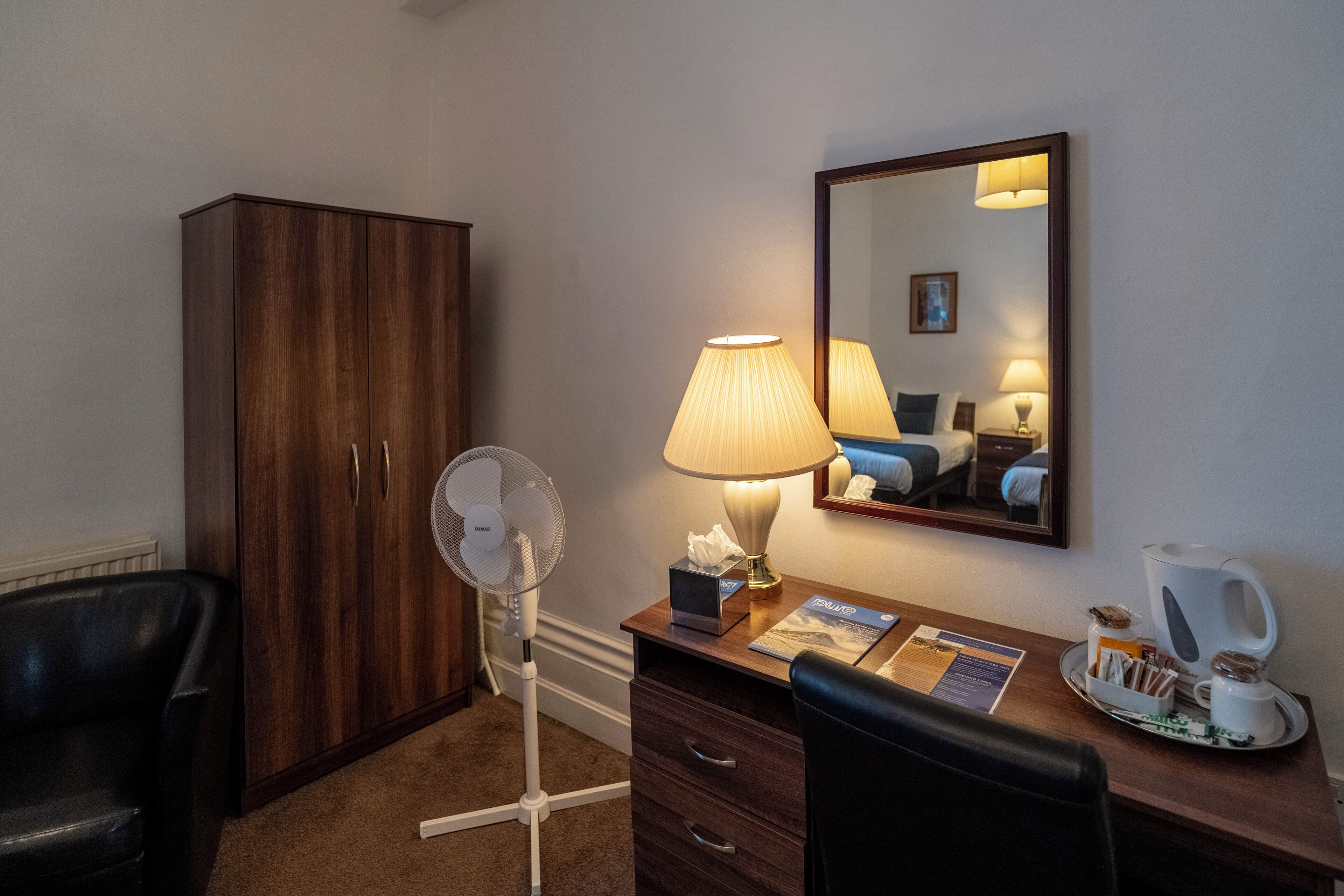 A hotel room with a wooden wardrobe, a desk with a lamp, a fan, a mirror, a coffee maker, and a chair.
