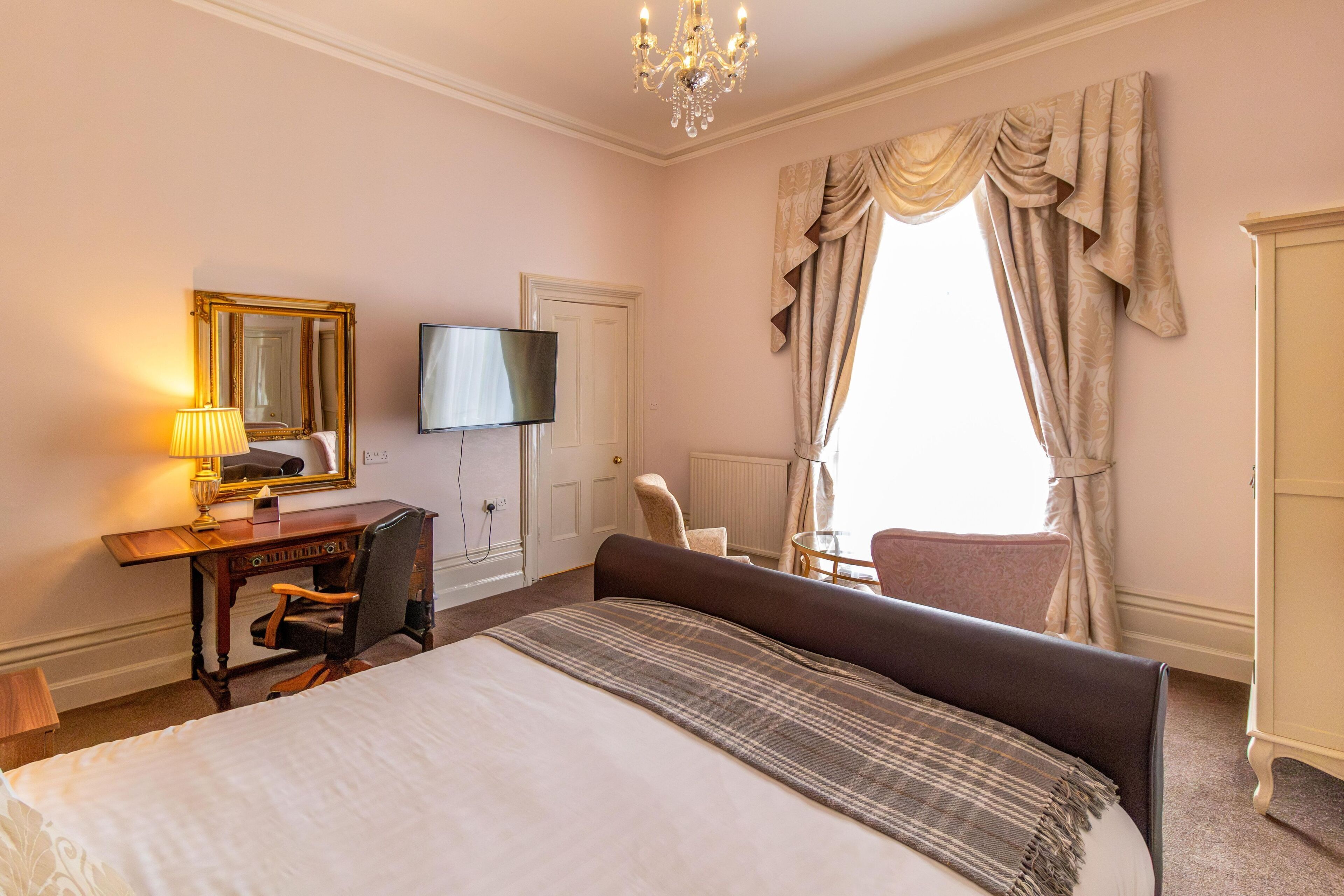 Una habitación de hotel con una cama grande, un escritorio con lámpara, televisor, una silla y una ventana con cortinas.