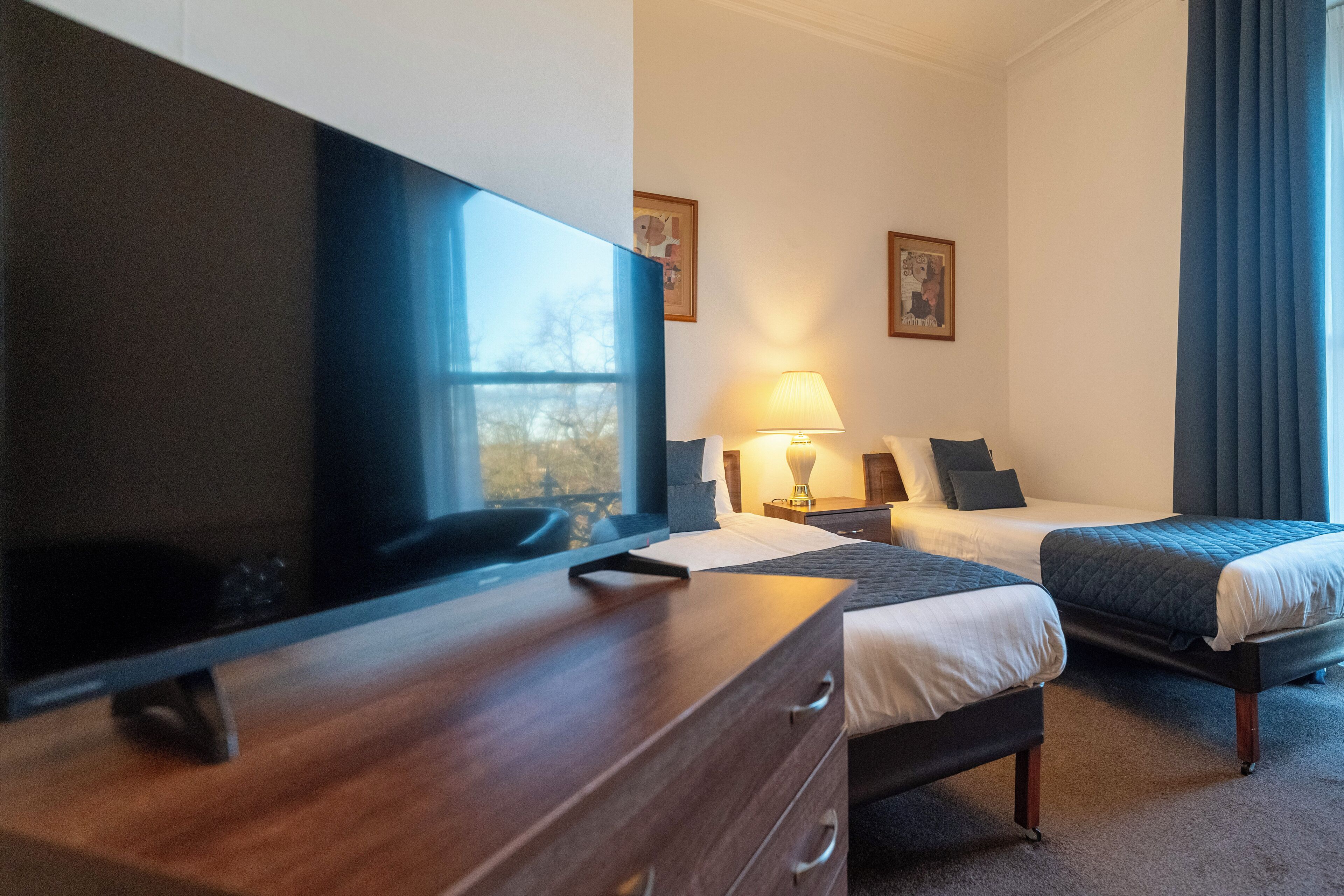 Une chambre d’hôtel dotée d’un grand téléviseur à écran plat, de deux lits avec une literie sombre, d’une table de chevet avec une lampe et d’une fenêtre avec des rideaux.