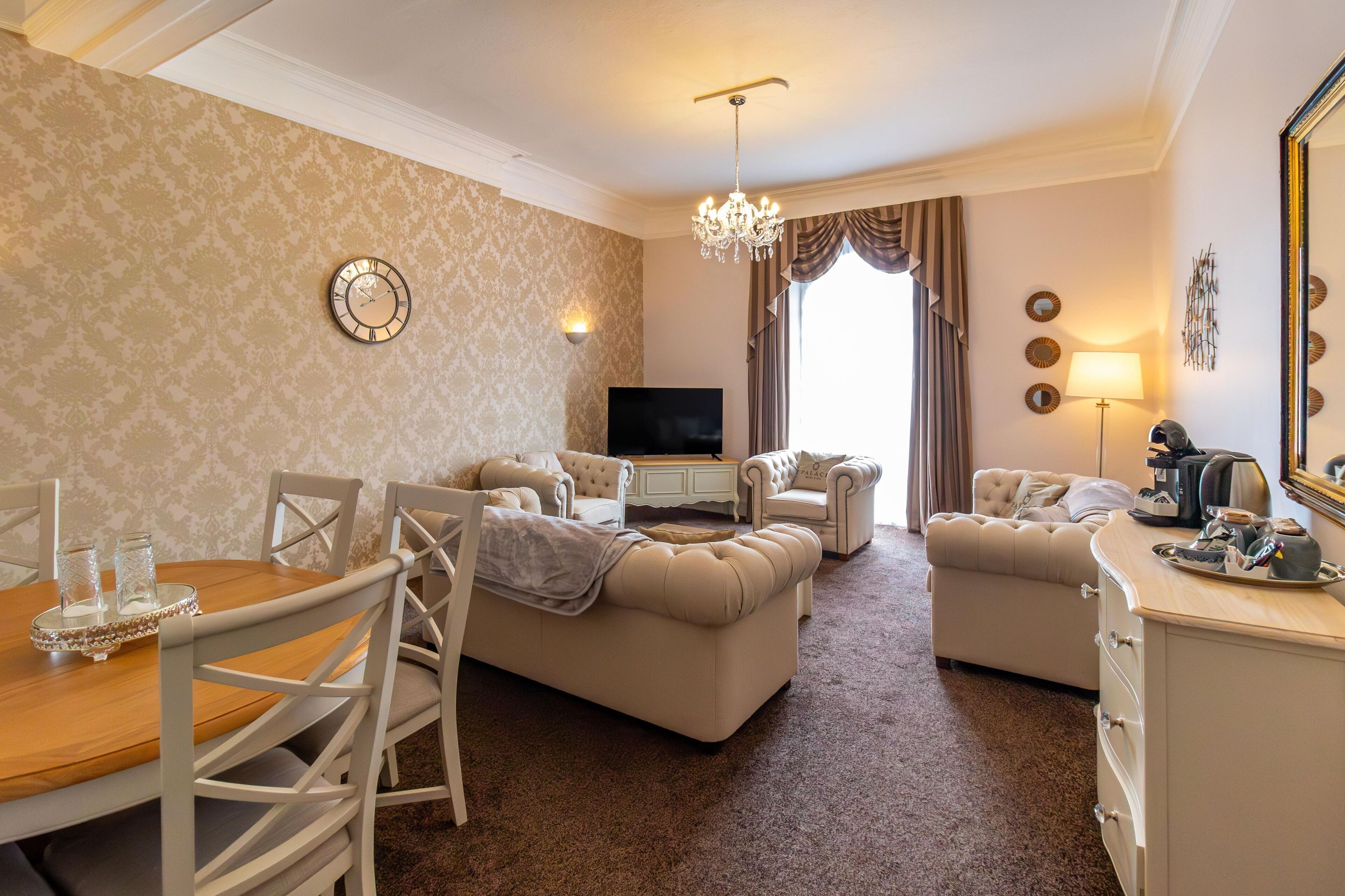 A well-appointed living room with a wooden dining table, beige sofas, a TV, and a chandelier.
