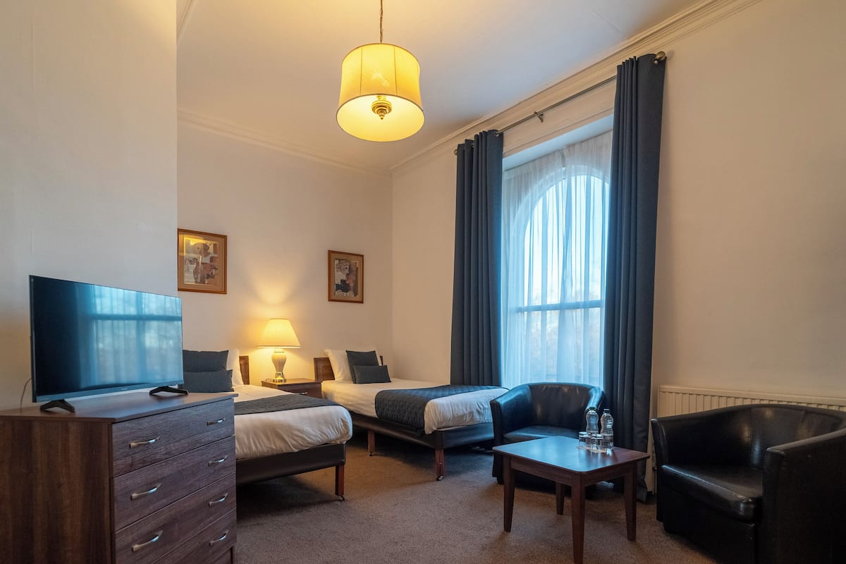 Ein Hotelzimmer mit zwei Einzelbetten, einem Schreibtisch, einem Stuhl, einem Fernseher und einer Lampe.