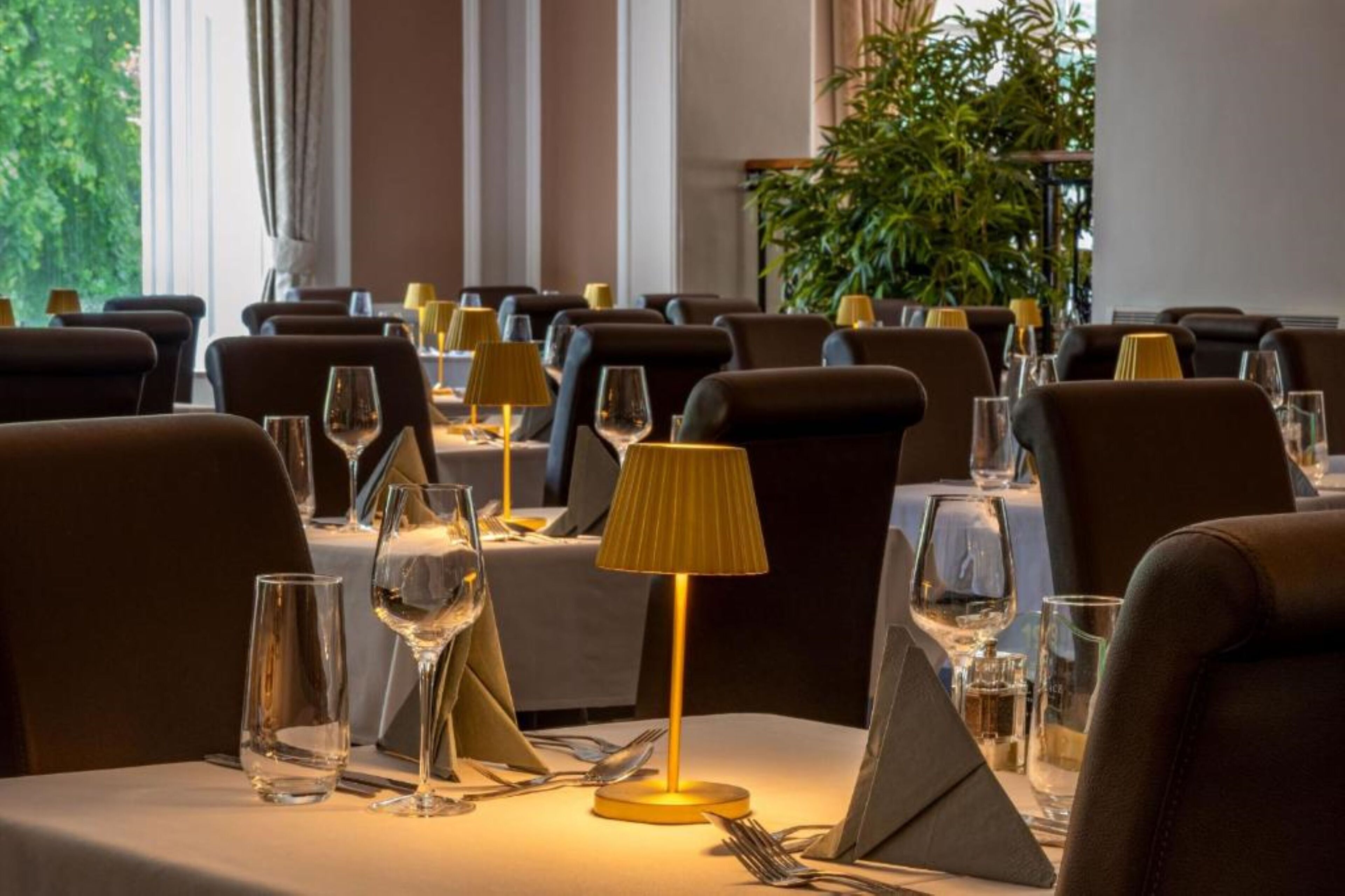Un restaurant où les tables sont dressées pour le service, offrant une ambiance chaleureuse et une lumière naturelle grâce à de grandes fenêtres.