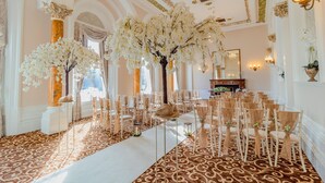 Un salón majestuoso con columnas decorativas, un arreglo floral central y filas de sillas.