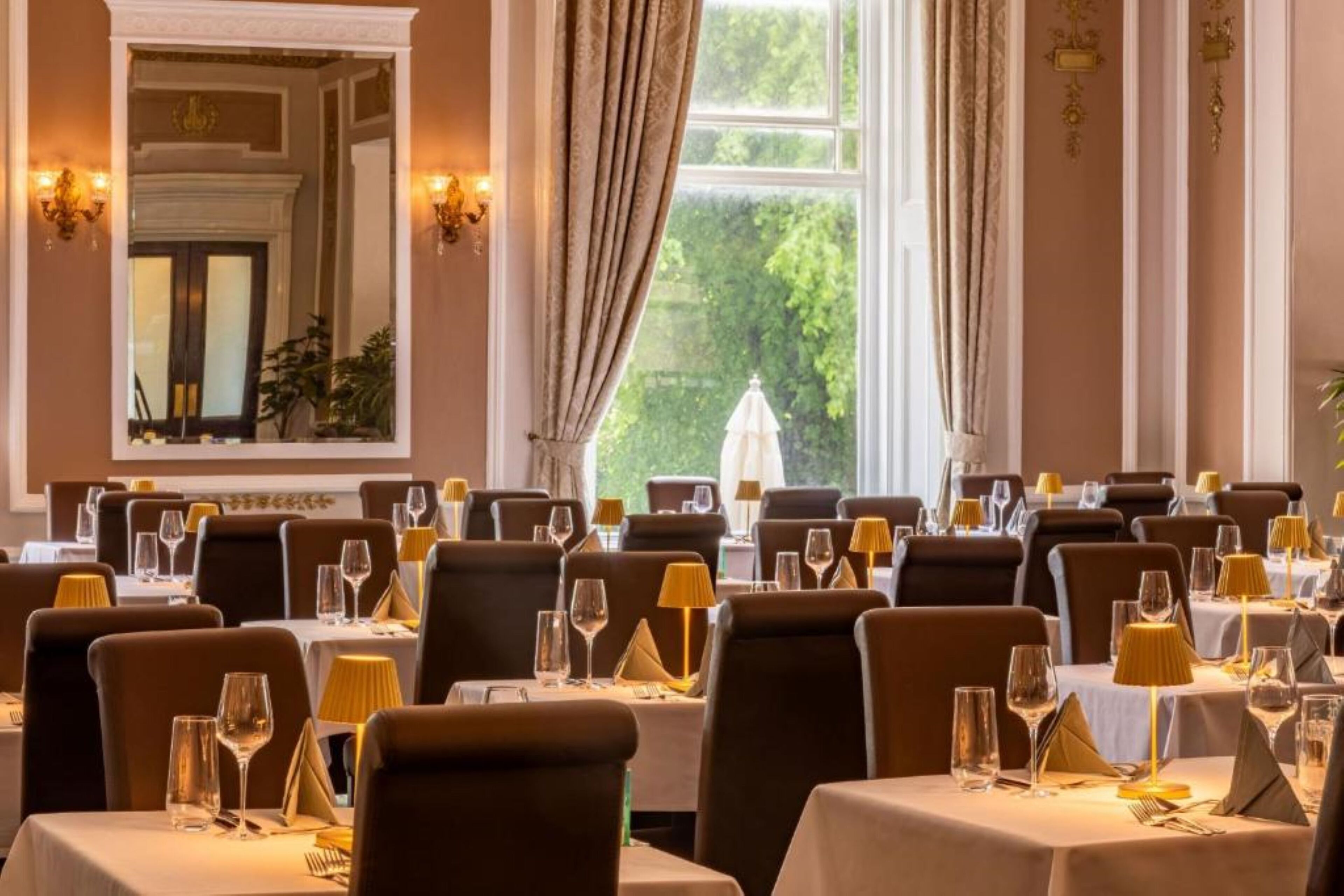 A dining area with tables set for service, a large mirror, and a view of greenery outside.