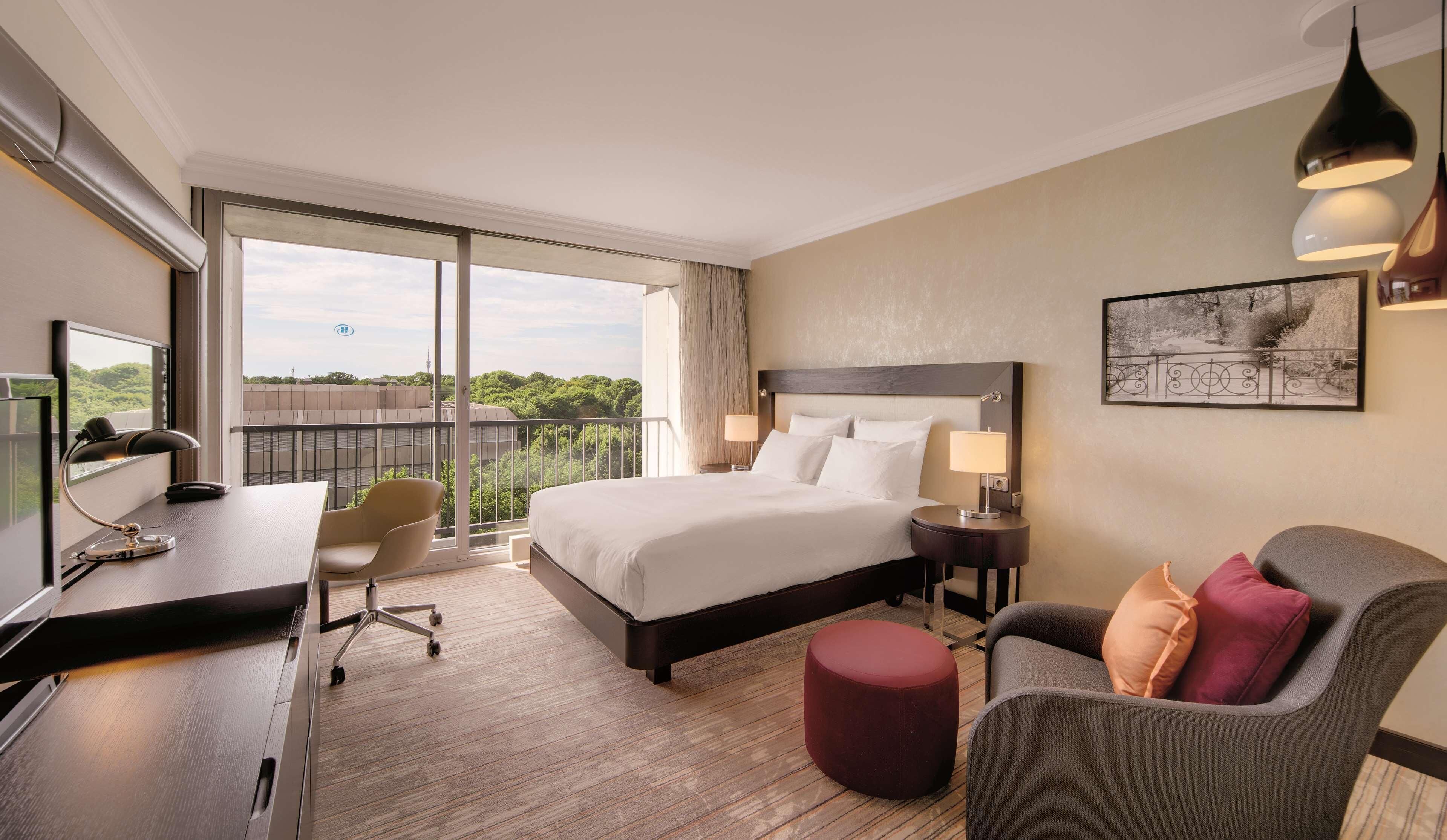 A hotel room with a large bed, a desk with a chair, a small ottoman, a nightstand with a lamp, and a view of greenery through the balcony.