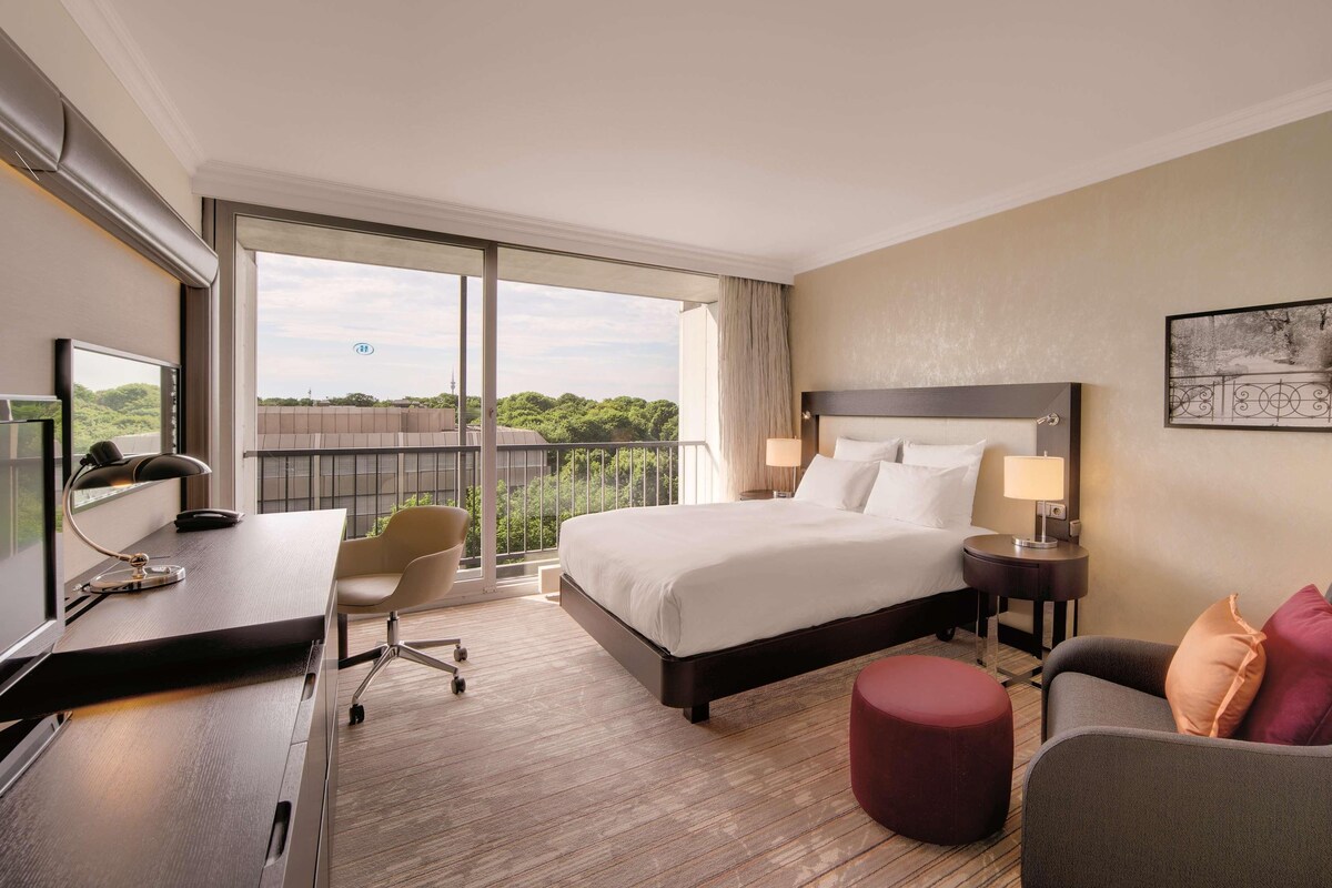 A hotel room with a large bed, a desk with a chair, a small ottoman, a nightstand with a lamp, and a view of greenery through the balcony.