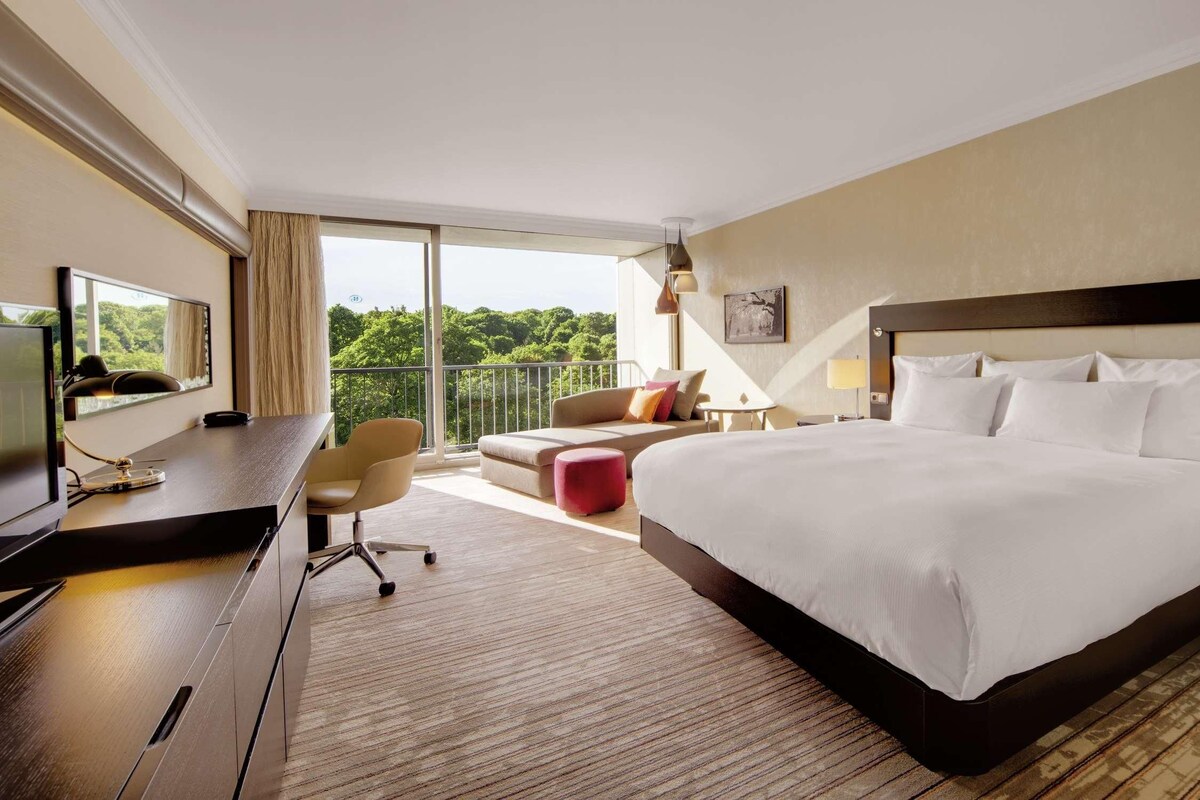 A hotel room with a large bed, a desk with a TV, a sofa, and a balcony with a view of greenery.