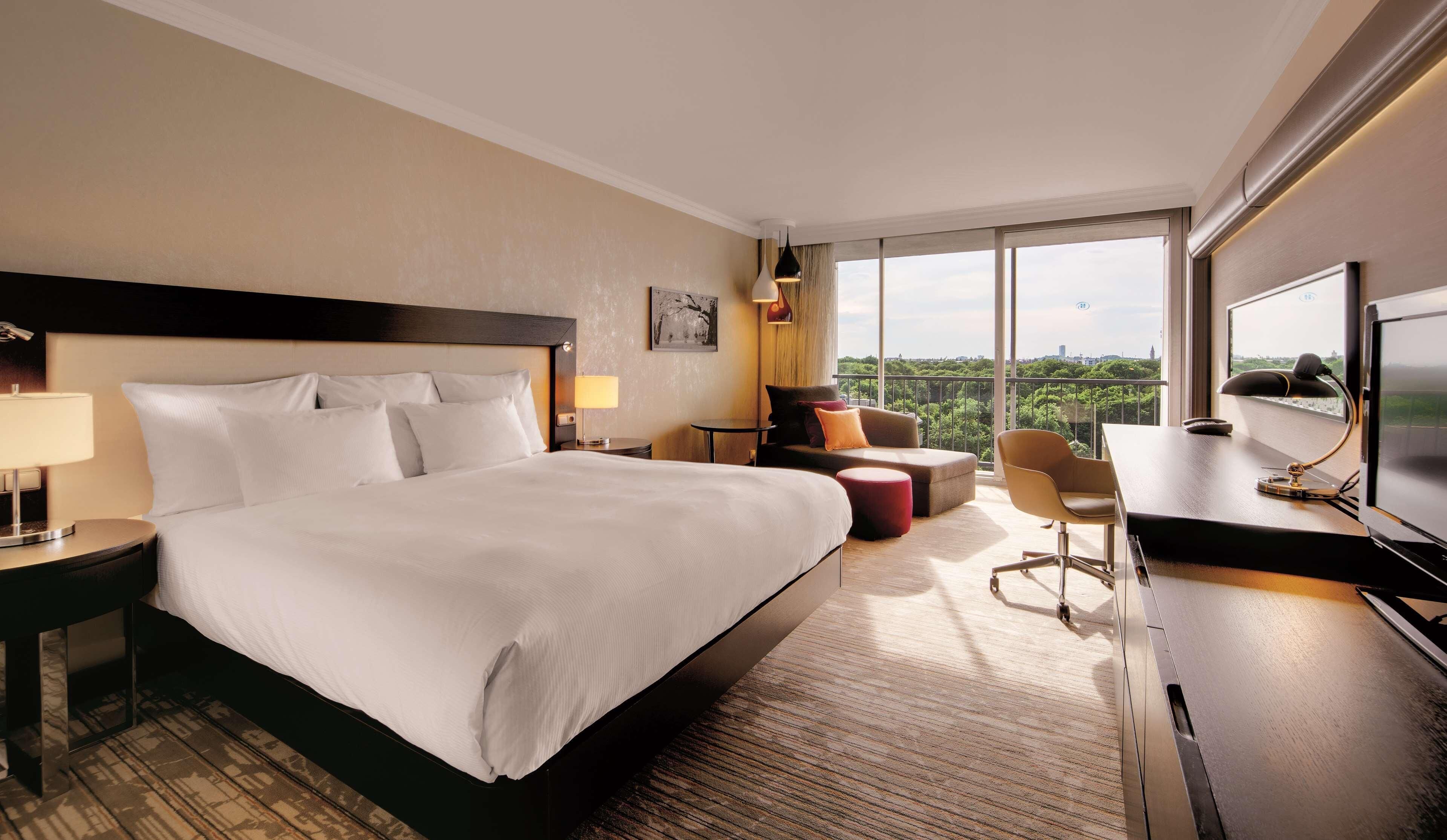 A hotel room with a large bed, a desk with a computer, a chair, a small table, and a TV.
