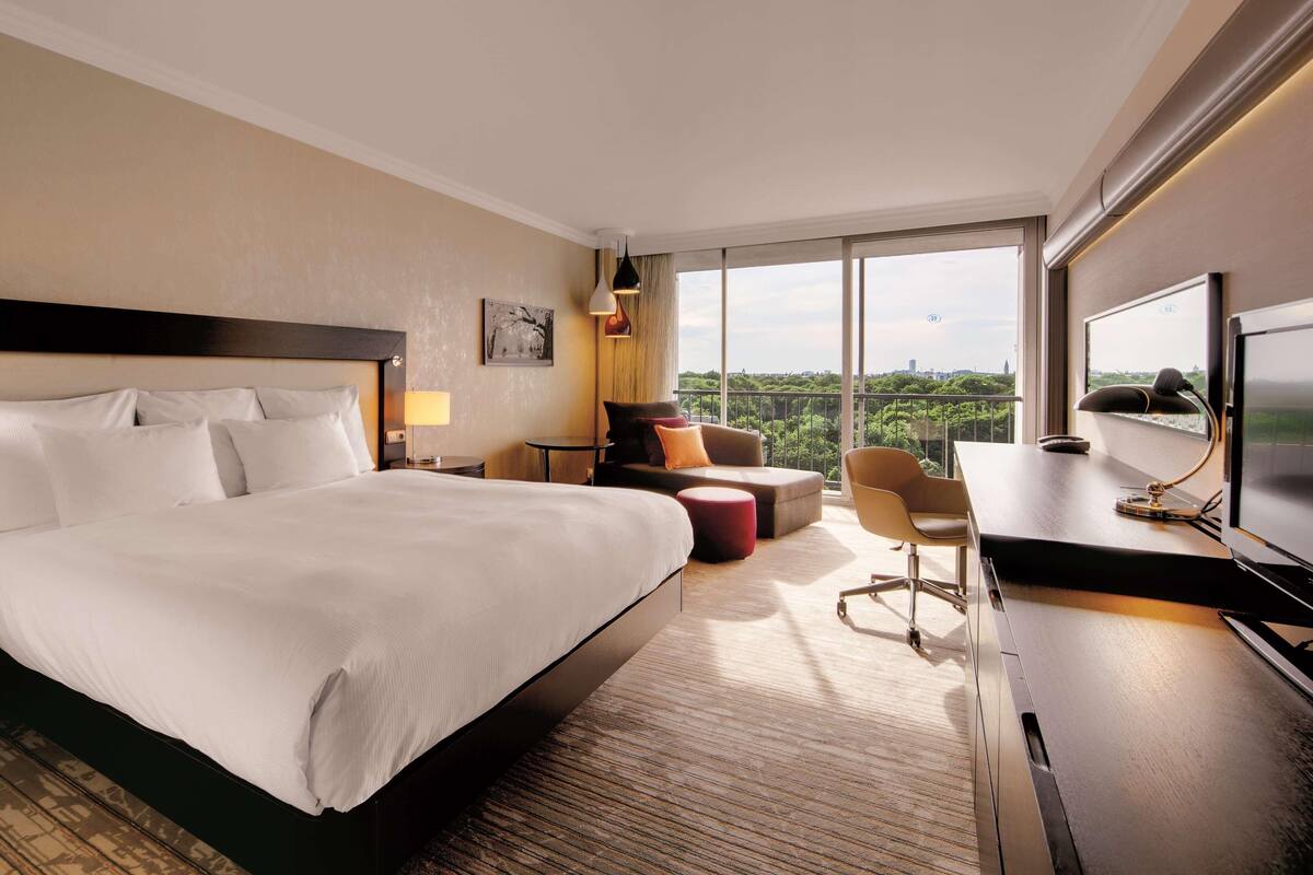 Ein Hotelzimmer mit einem großen Bett, einem Schreibtisch mit Computer, einem Sessel, einem kleinen Tisch und einem Fernseher.