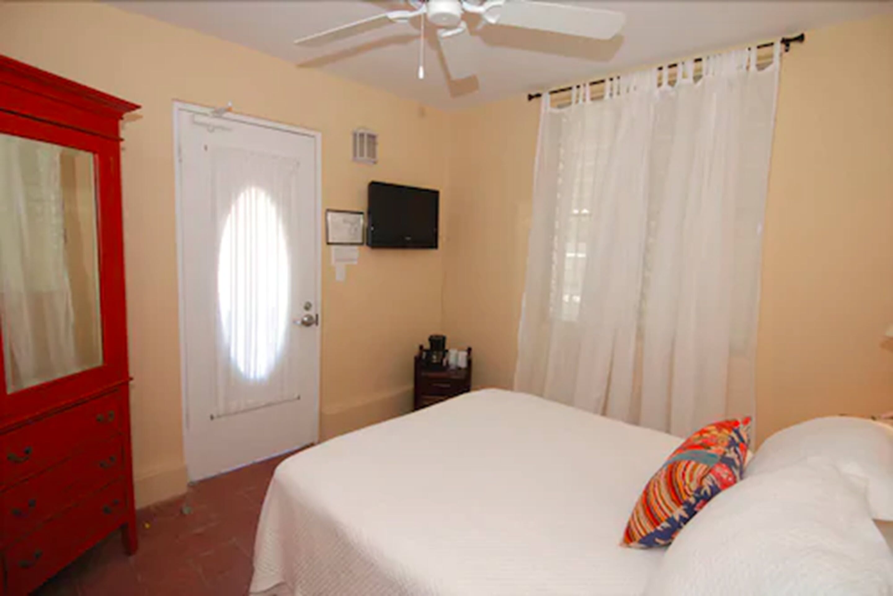 A bedroom with a bed, a red dresser, a ceiling fan, a TV, and a window with curtains.