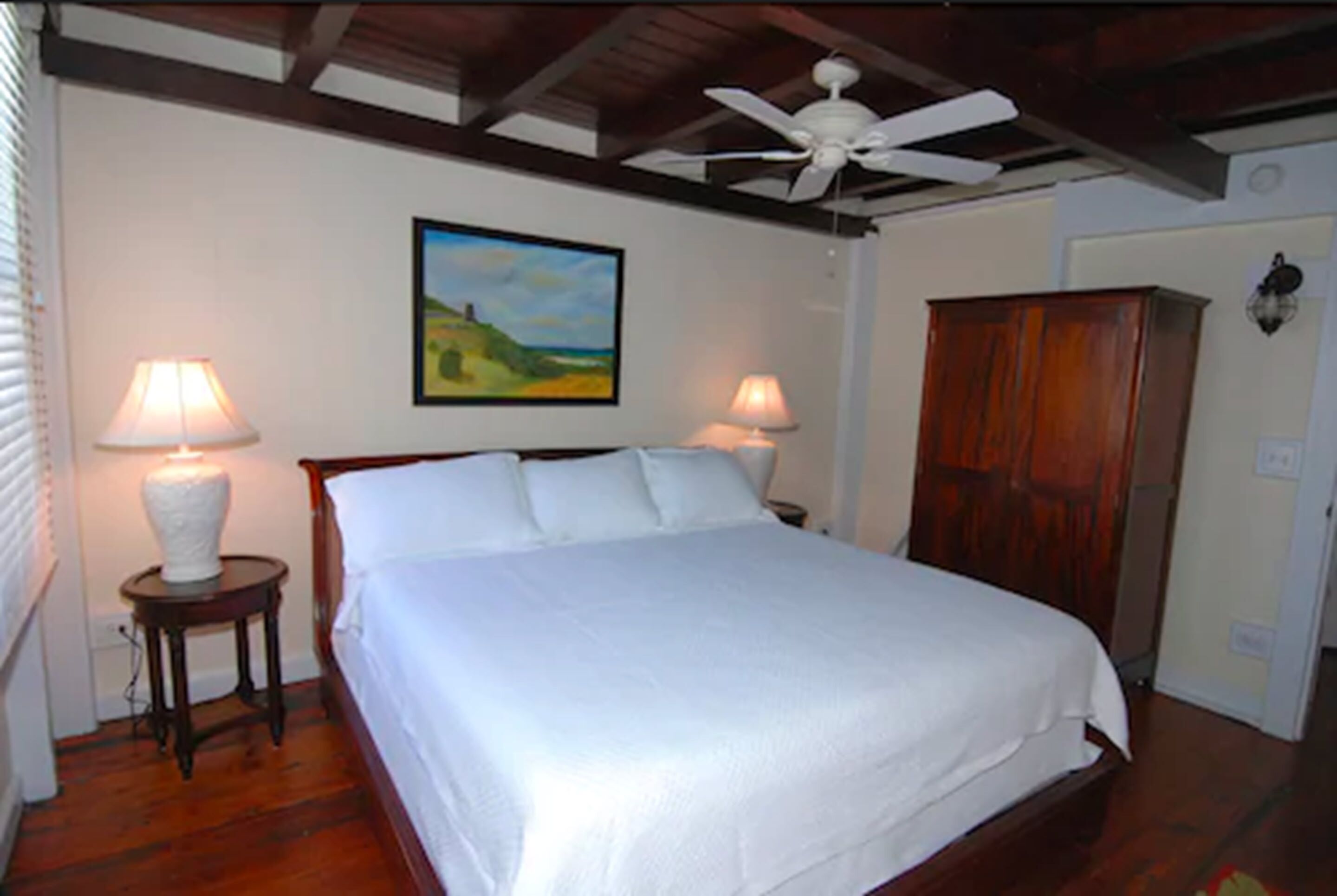 A bedroom with a bed, two bedside lamps, a ceiling fan, a wooden wardrobe, and a painting on the wall.