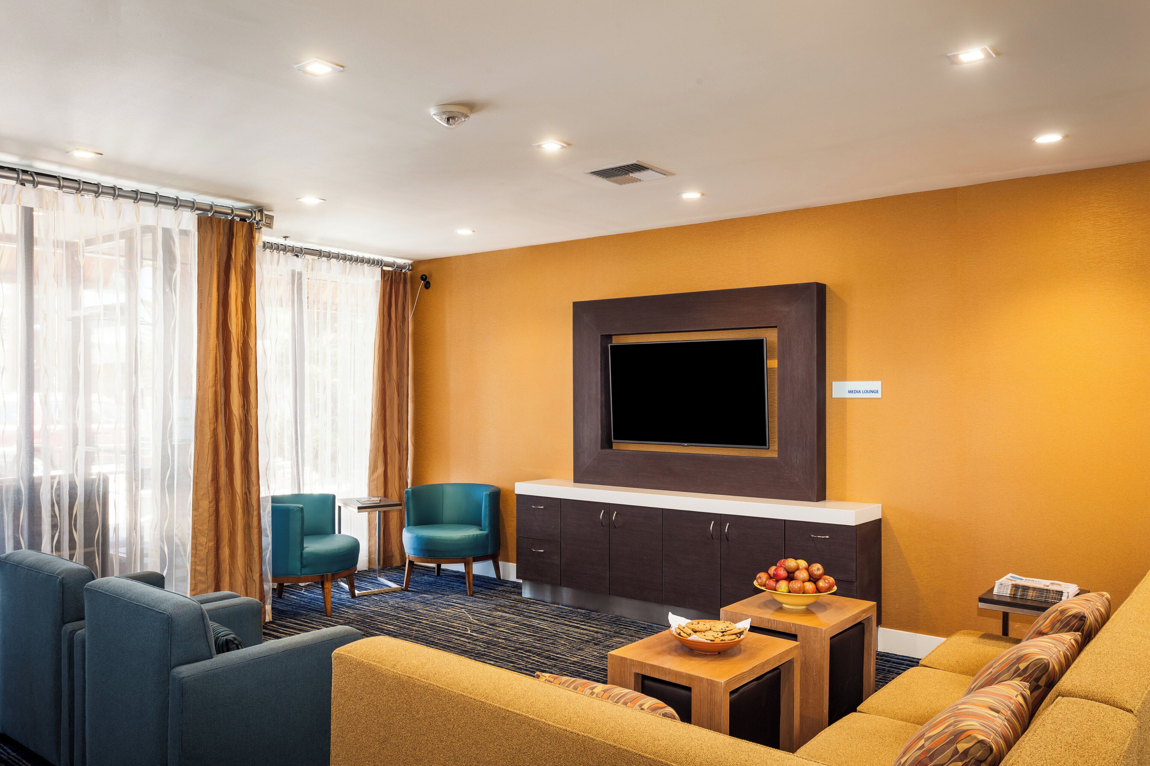 A modern living room with a flat-screen TV, a sofa, two armchairs, a coffee table, and a small table with apples and a bowl.