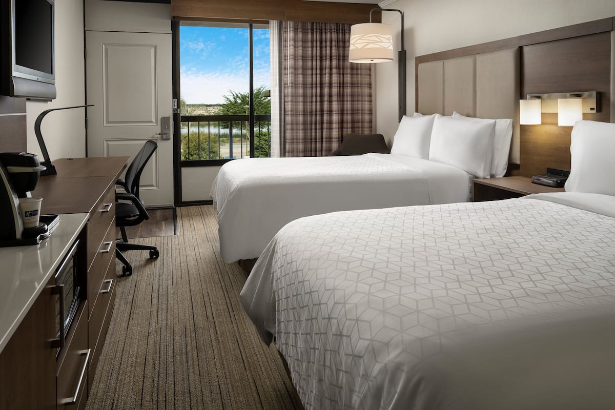 A hotel room with two beds, a desk with a coffee maker, a TV, and a view of the outdoors.