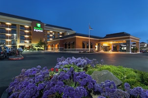 Un hotel Holiday Inn Express con estacionamiento y una zona ajardinada con flores moradas.