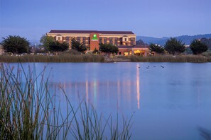 A Holiday Inn Express hotel with a large building, a body of water in the foreground, and trees around. - Holiday Inn Express at Monterey Bay by IHG (Seaside)