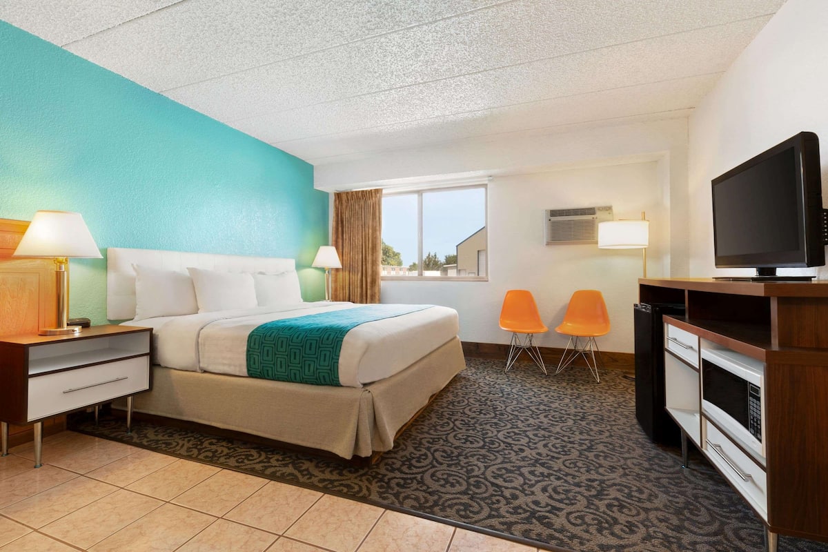 A hotel room with a bed, a desk with a television, two chairs, and a window with curtains.