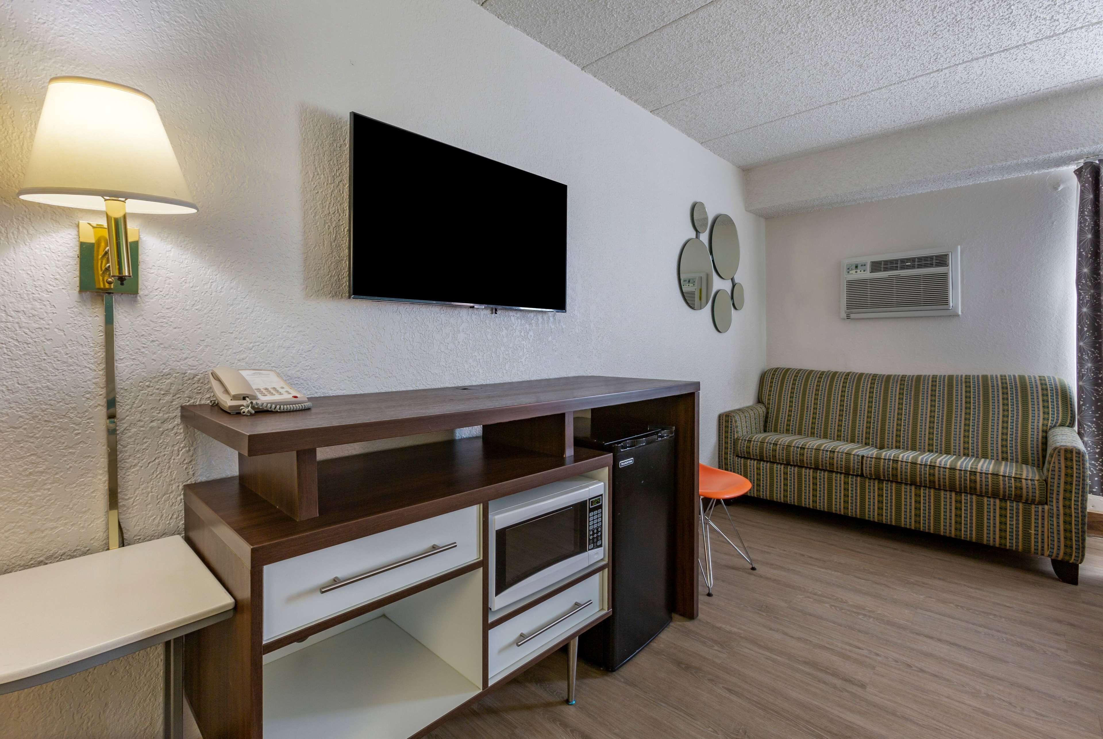 A modern living room with a flat-screen TV, a desk with a lamp, a microwave, a refrigerator, and a striped sofa.