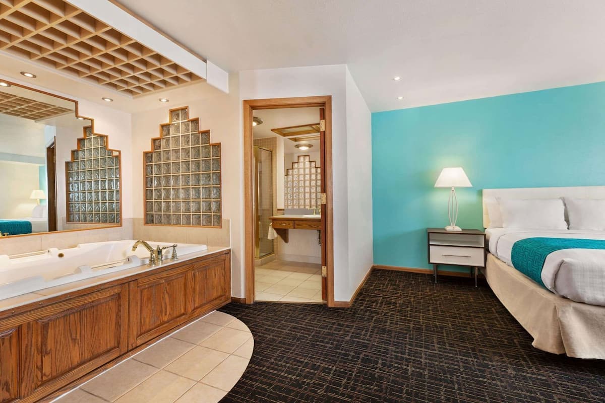 A hotel room with a large wooden vanity, a separate shower area, a bed with a teal accent wall, and a nightstand with a lamp.