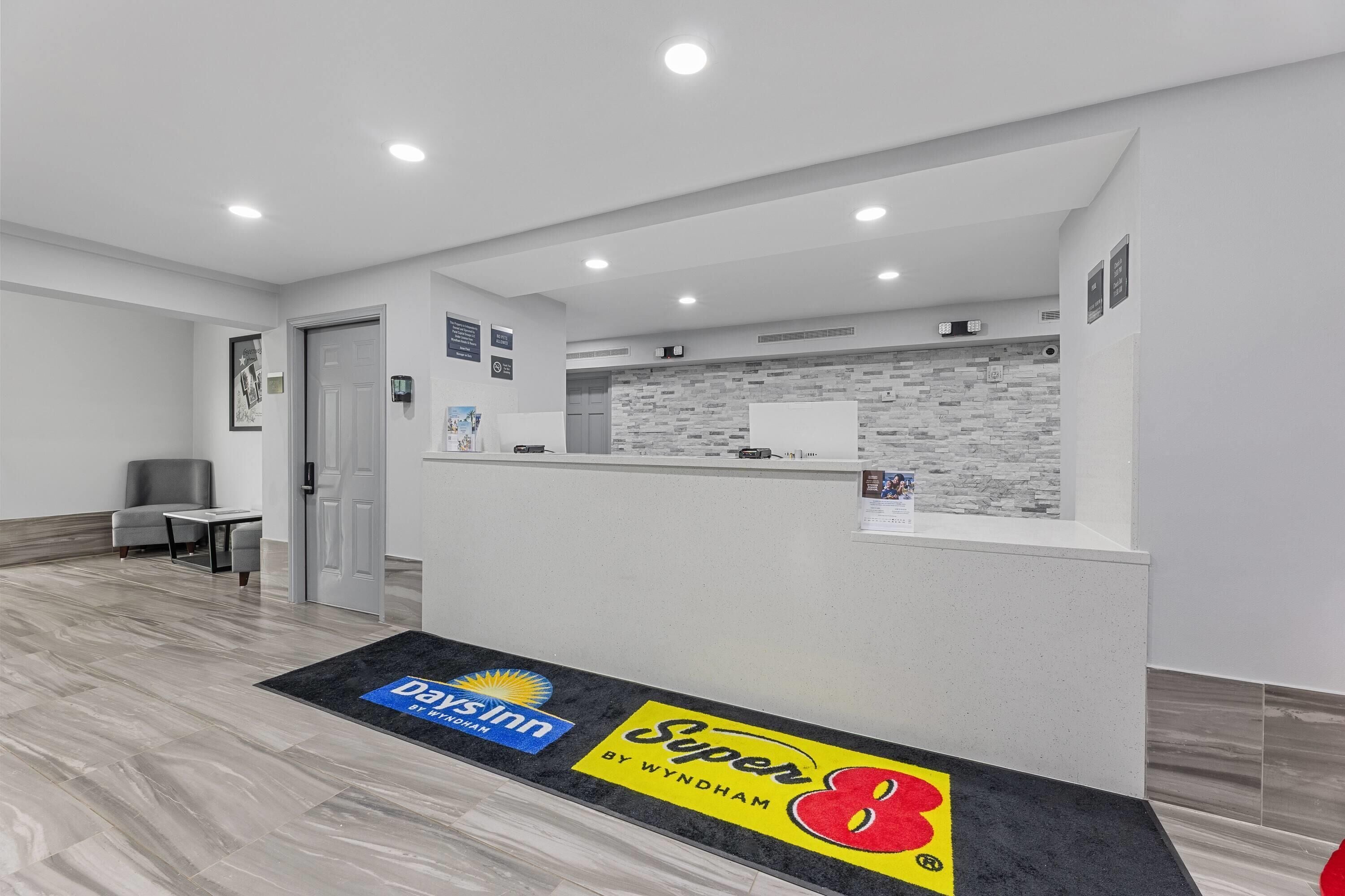 A hotel reception area with a reception desk, a waiting area with a chair and table, and a reception desk with a logo for 'Super 8 by Wyndham'.