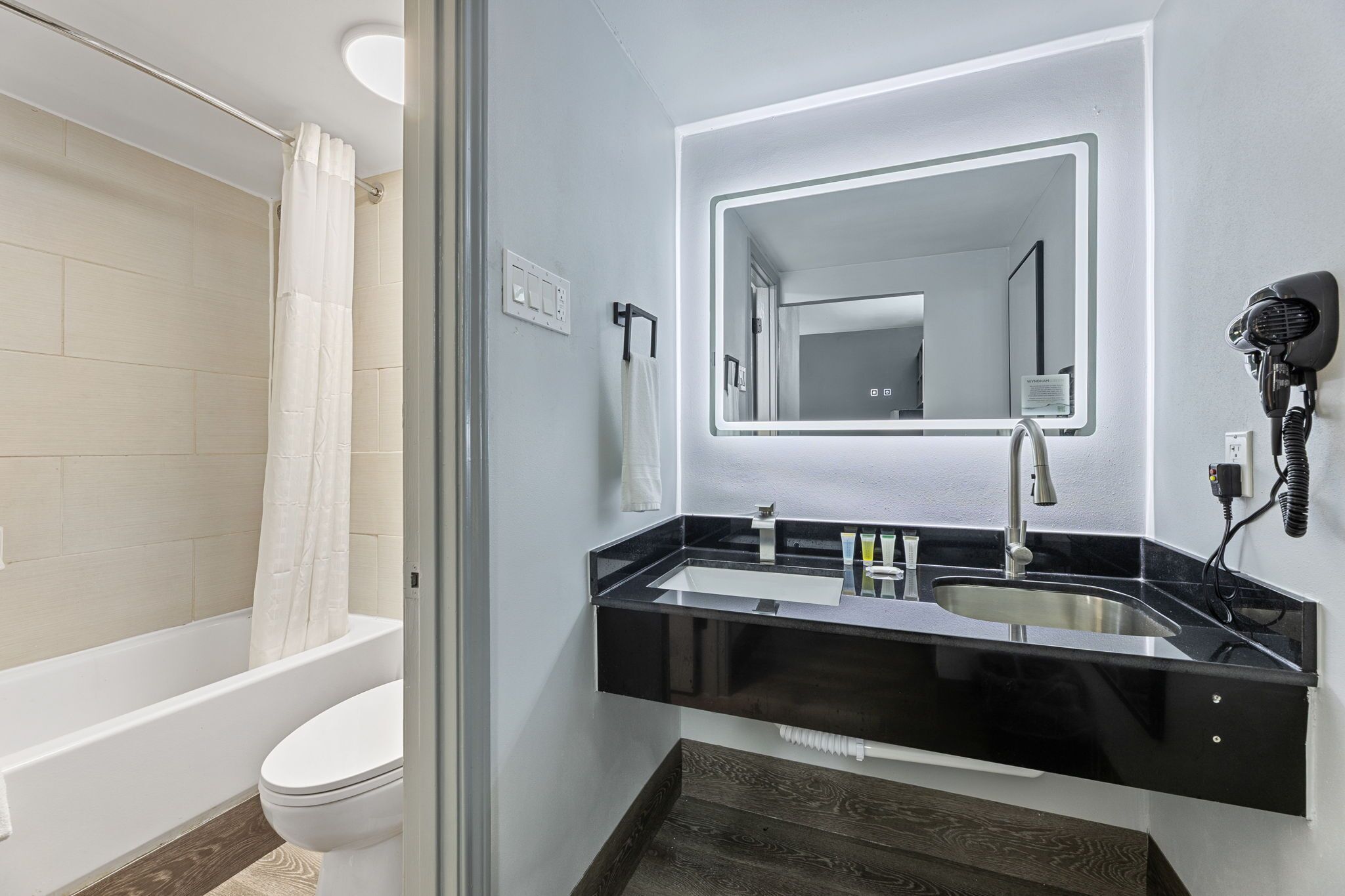 A modern bathroom with a bathtub, a toilet, a sink with a black countertop, and a hairdryer mounted on the wall.