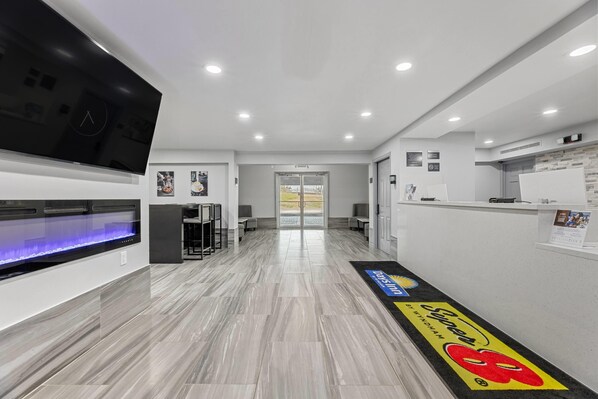 A modern hotel lobby with a reception desk, a flat-screen TV, a blue-lit aquarium, and a reception area with a logo mat.