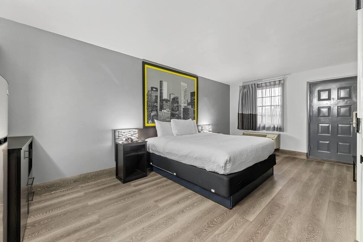 Une chambre moderne avec un lit, une table de chevet, un tableau accroché au mur, une porte et une fenêtre avec des rideaux.