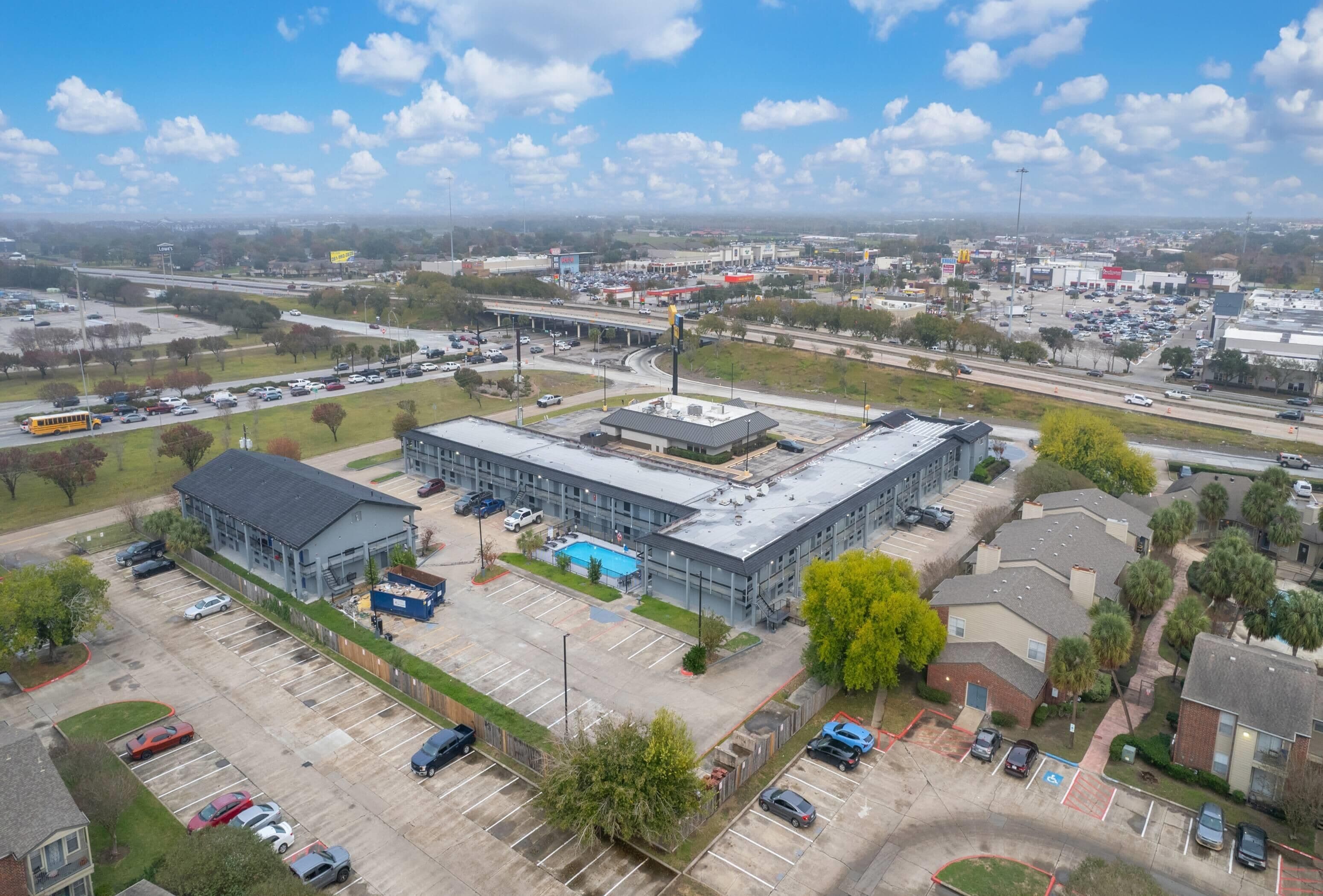 Vista aérea de un edificio comercial con estacionamiento, junto a una autopista y una zona residencial.