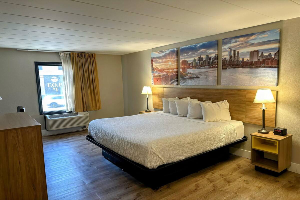 A hotel room with a large bed, two bedside lamps, a nightstand, a window with curtains, and a cityscape painting on the wall.