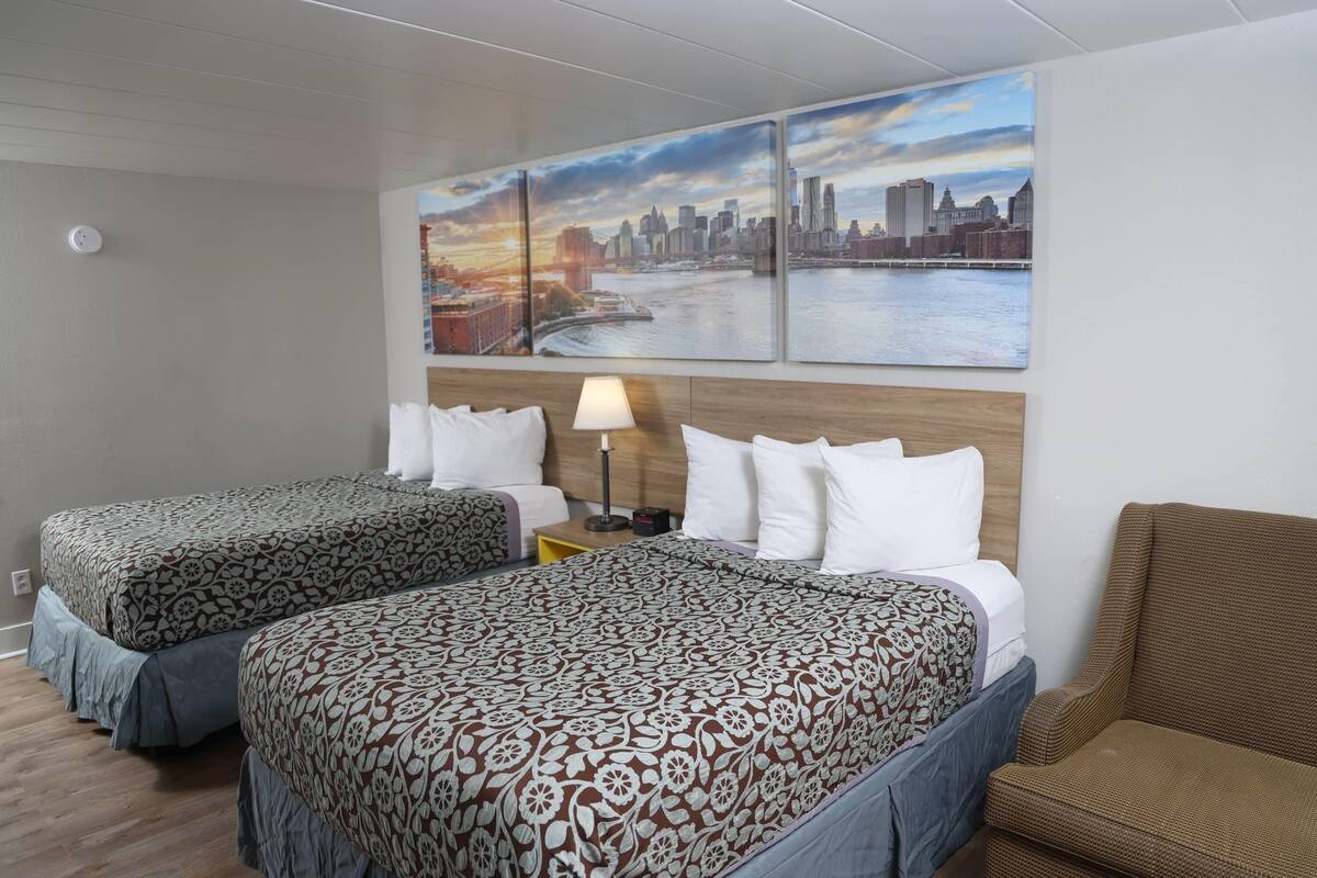 A hotel room with two beds, a cityscape painting, a lamp, and a wooden headboard.
