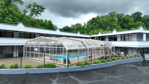 Une piscine couverte, protégée par une structure en verre, entourée d’une clôture, et un espace patio aménagé avec des sièges d’extérieur.