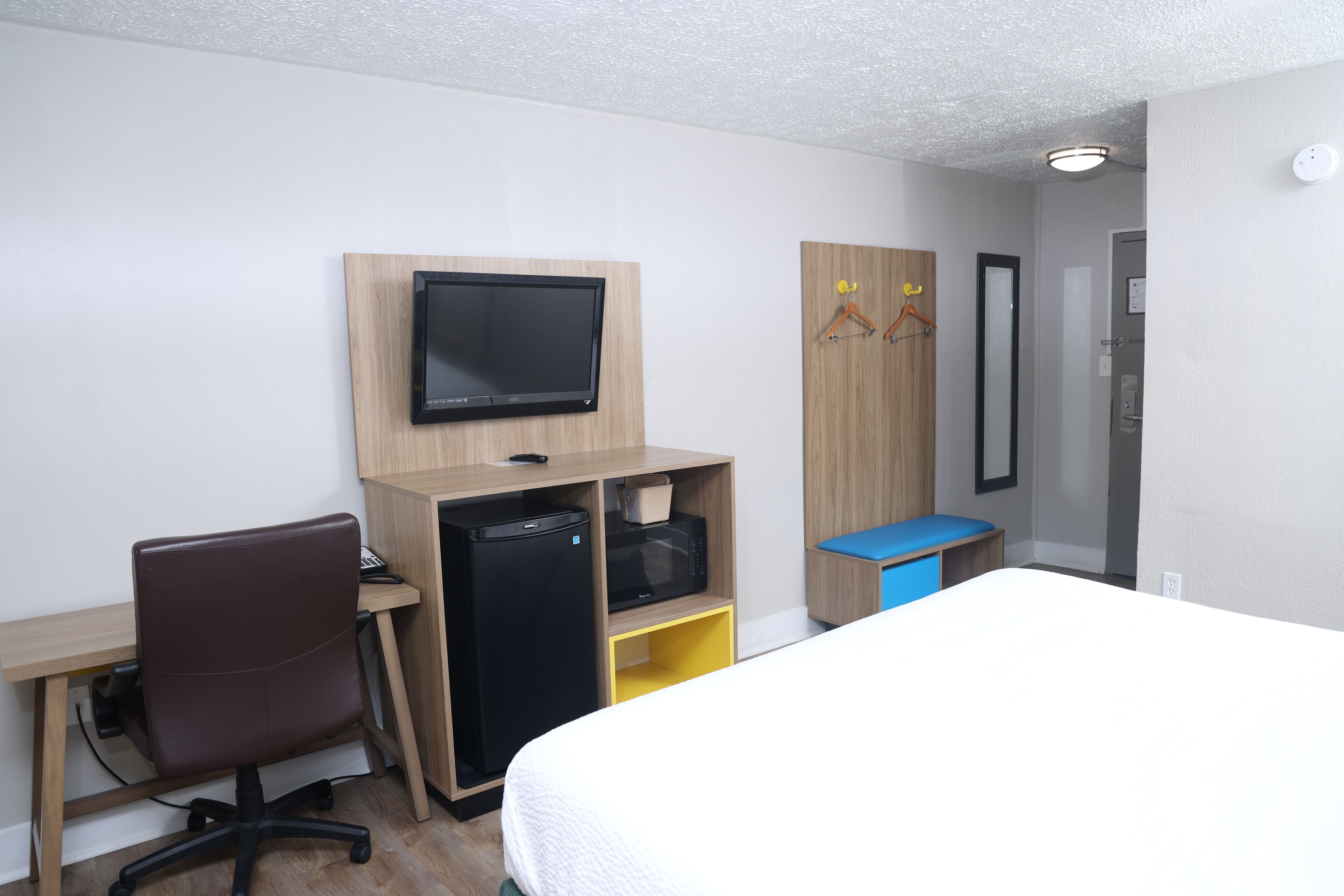 Habitación de hotel con cama, escritorio con computadora, refrigerador y televisor montado en la pared.