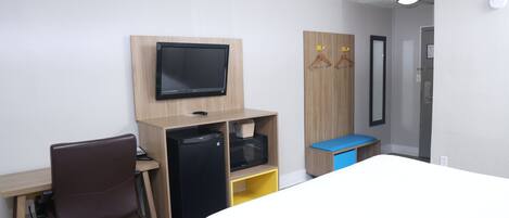 Habitación de hotel con cama, escritorio con computadora, refrigerador y televisor montado en la pared.