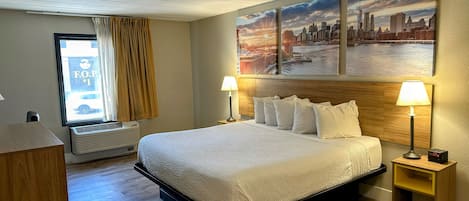 Habitación de hotel con cama, dos lámparas de noche, una ventana con cortinas, un aire acondicionado de pared y un cuadro de paisaje urbano sobre la cama.