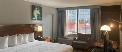 A hotel room with a large bed, a desk, a chair, a lamp, and a view of the outside.