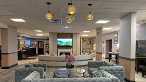 A hotel lobby with a seating area featuring tufted sofas, a round coffee table, and decorative lighting. A flat-screen TV is mounted on the wall, and a fireplace is visible in the background.