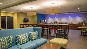 A modern hotel lobby with a blue sofa, a wooden coffee table, a bar area with stools, and a stone wall backdrop.