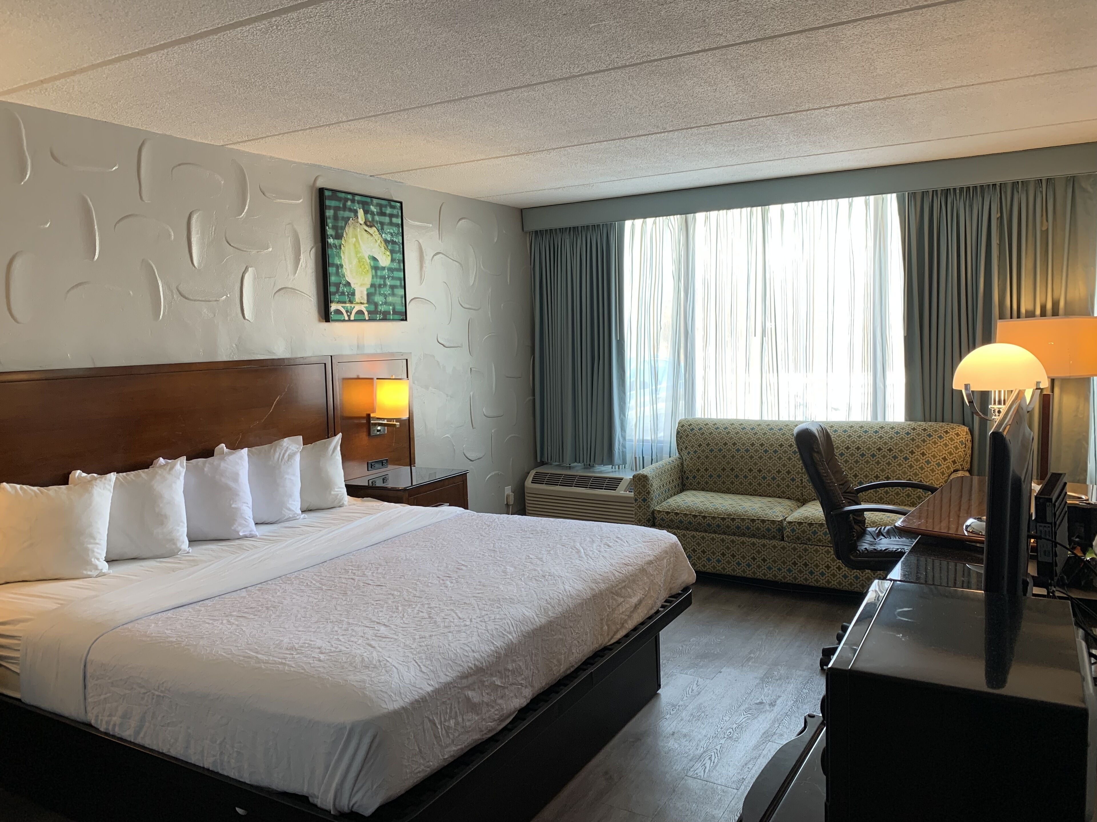 A hotel room with a large bed, a sofa, a desk with a lamp, and a window with curtains.