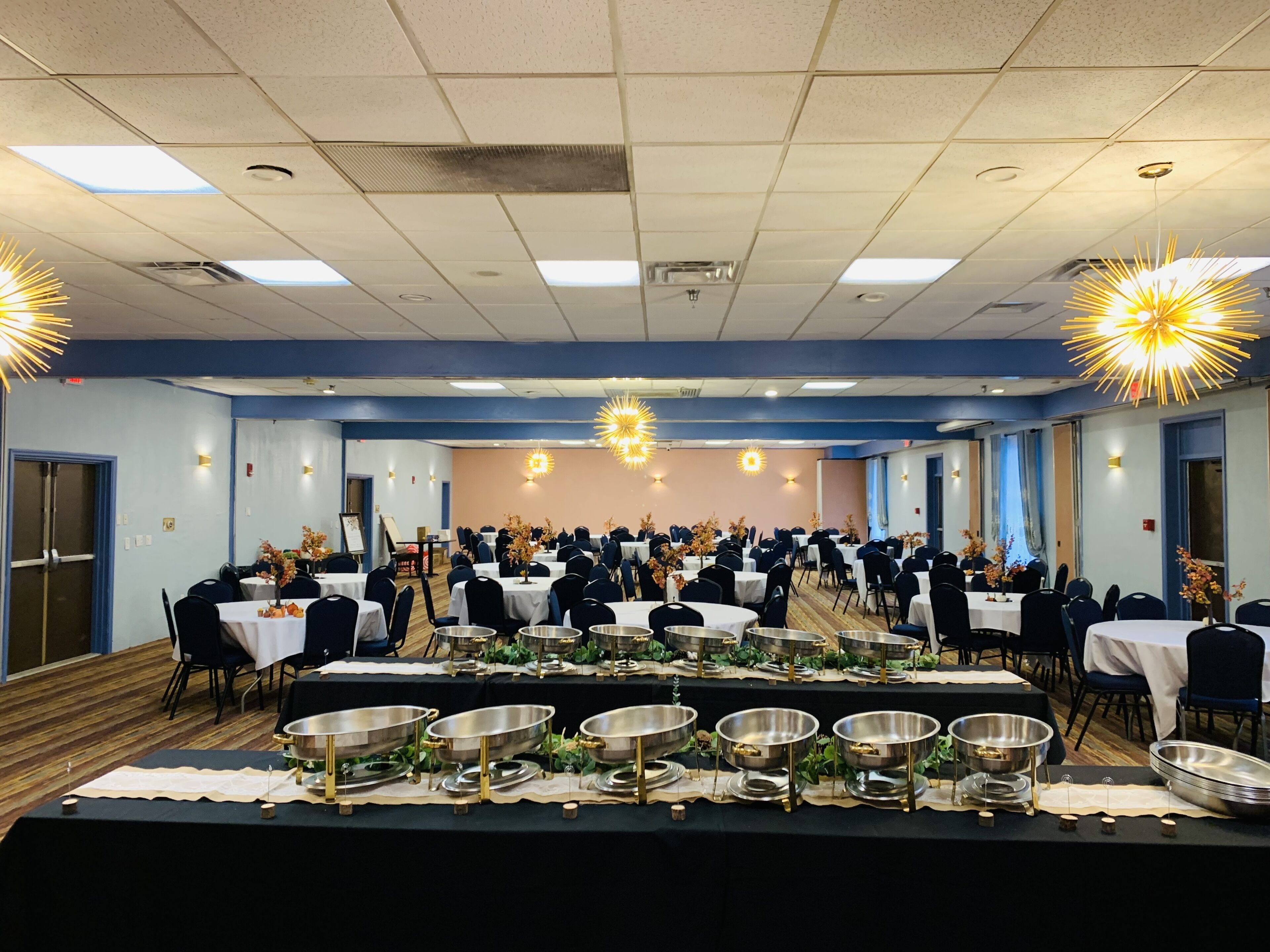 A banquet hall with multiple round tables, each with a white tablecloth and blue chairs. There are large, decorative chandeliers hanging from the ceiling, and the room has a high ceiling with visible air conditioning vents.