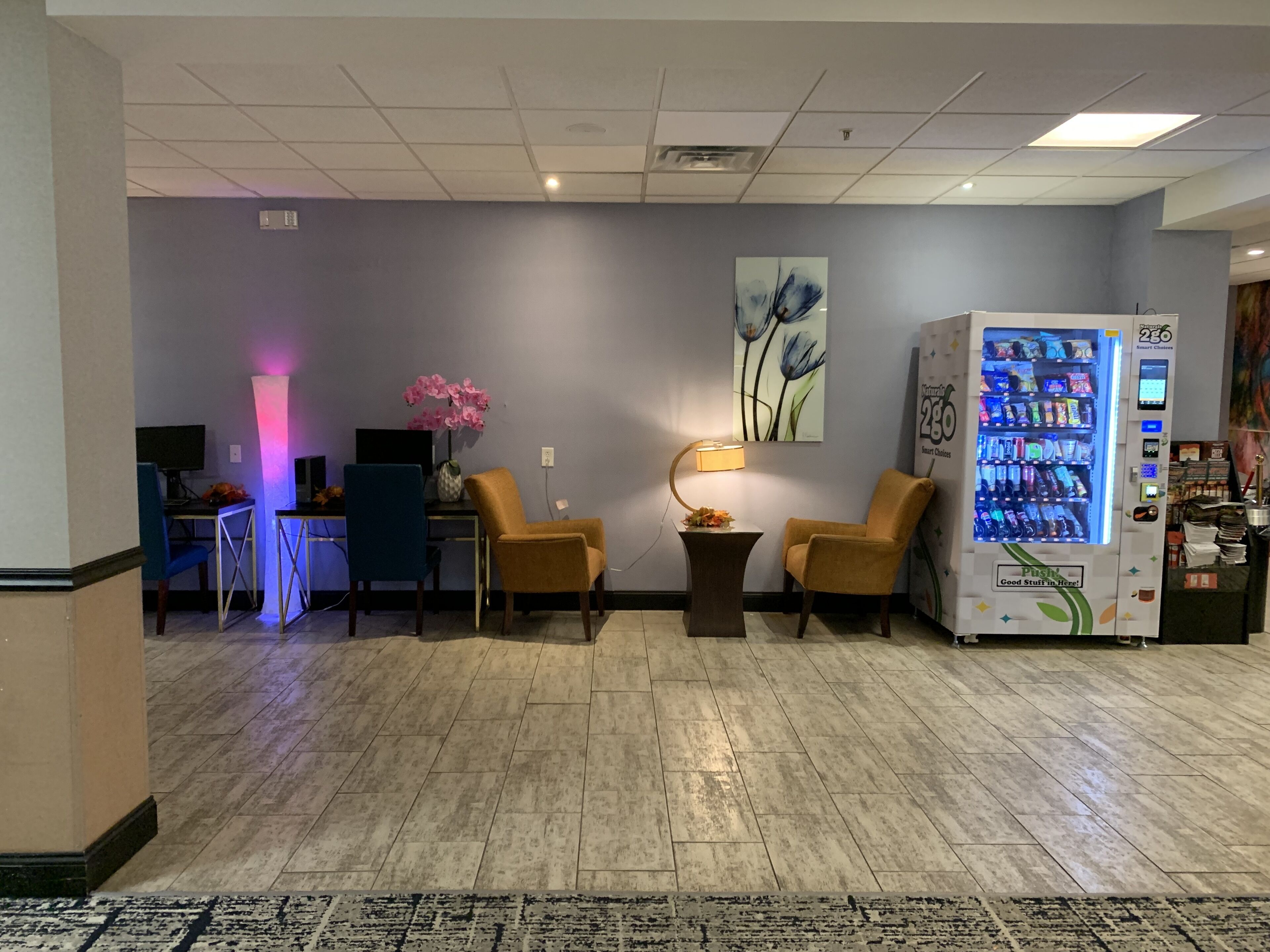 A lobby with a reception desk, two armchairs, a lamp, a wall-mounted painting, and a vending machine.