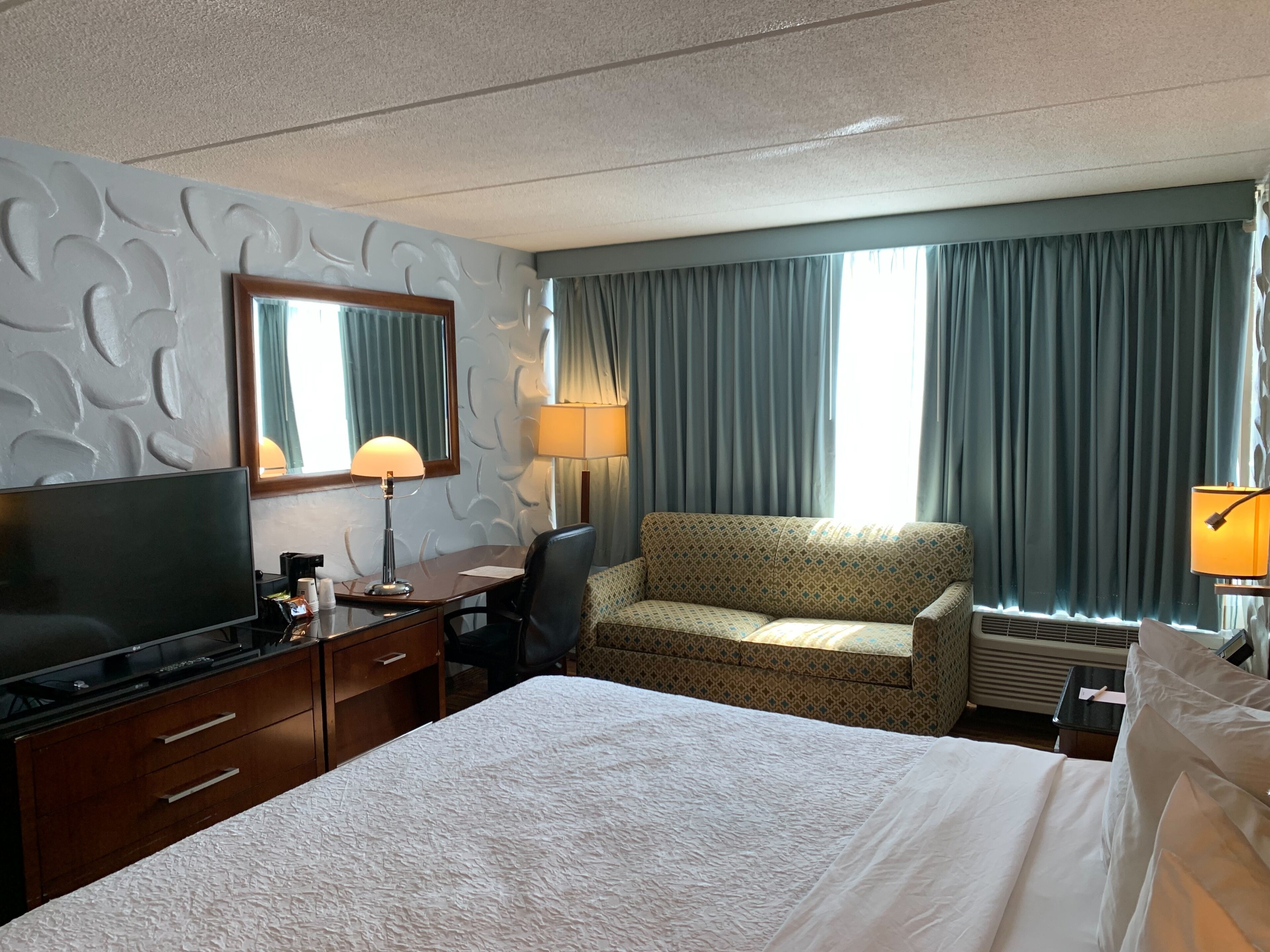 Habitación de hotel con cama, escritorio con televisor, silla, sofá, lámpara y ventana con cortinas.