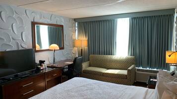 Habitación de hotel con cama, escritorio con televisor, silla, sofá, lámpara y ventana con cortinas.