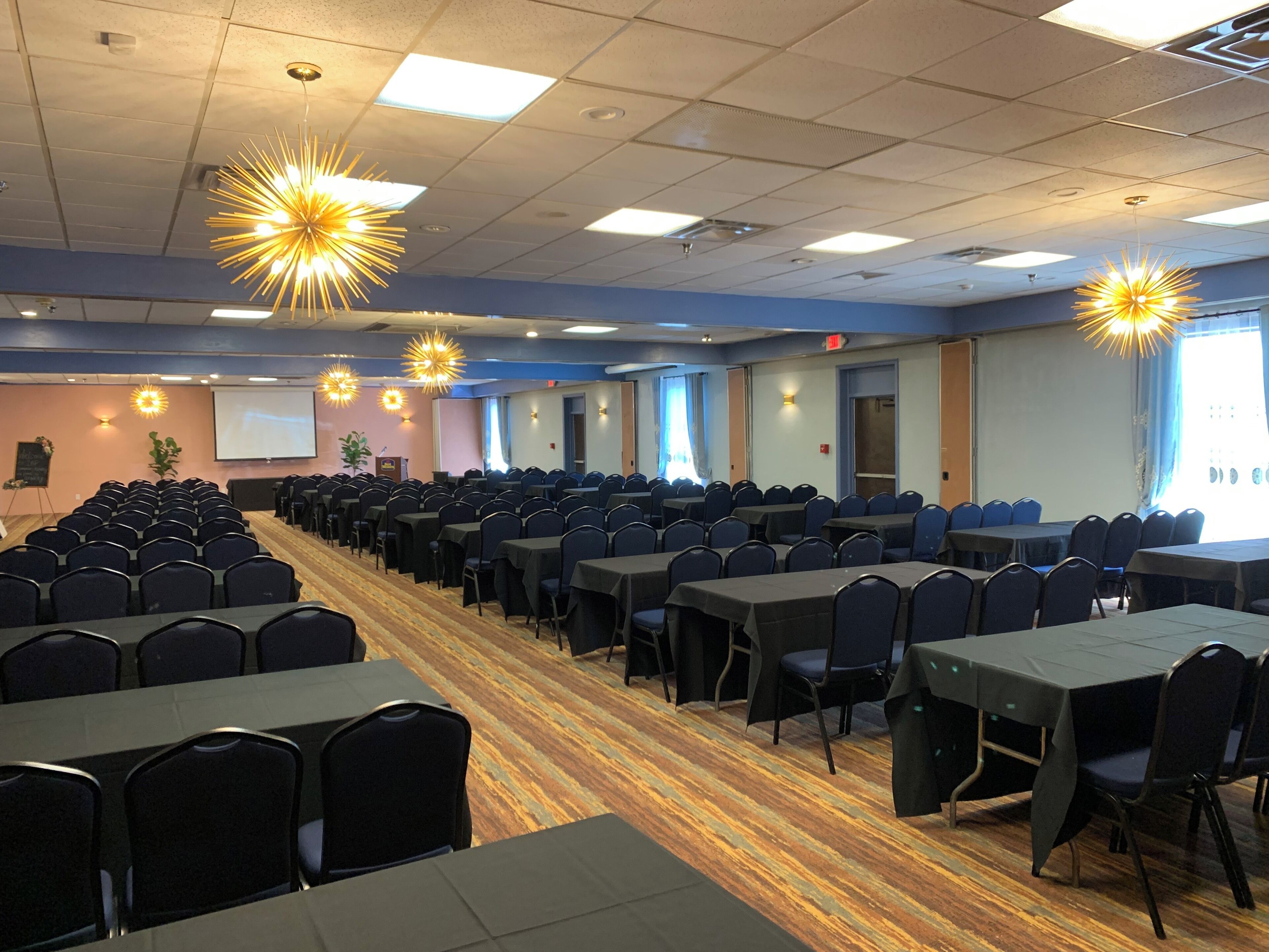 A conference room with rows of chairs and tables, ceiling lights, and a projector screen.