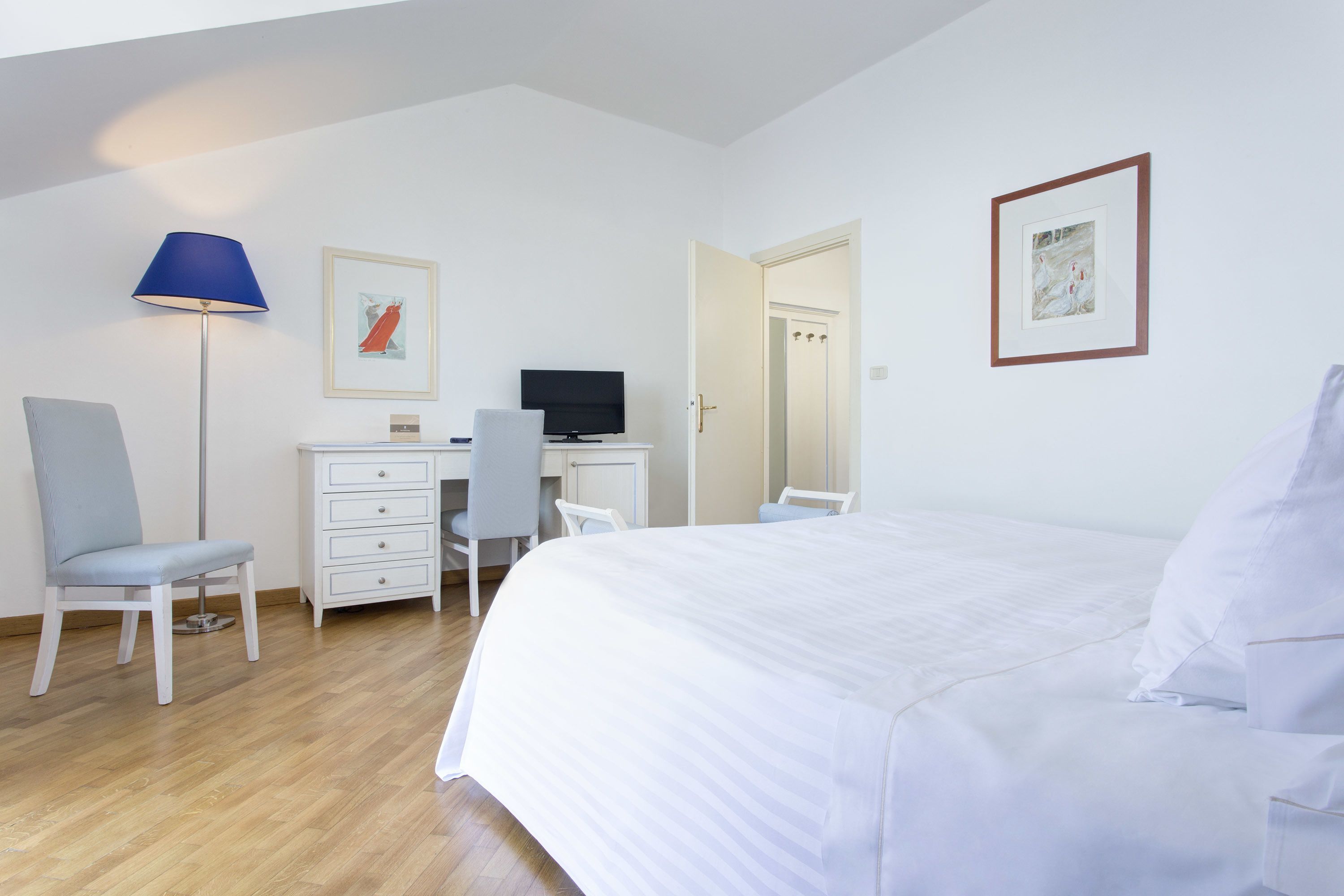 A hotel room with a bed, a desk with a computer, a chair, a lamp, and artwork on the walls.