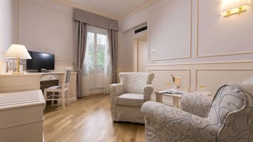 A hotel room with a wooden floor, a TV, a desk with a lamp, a chair, and a sofa.